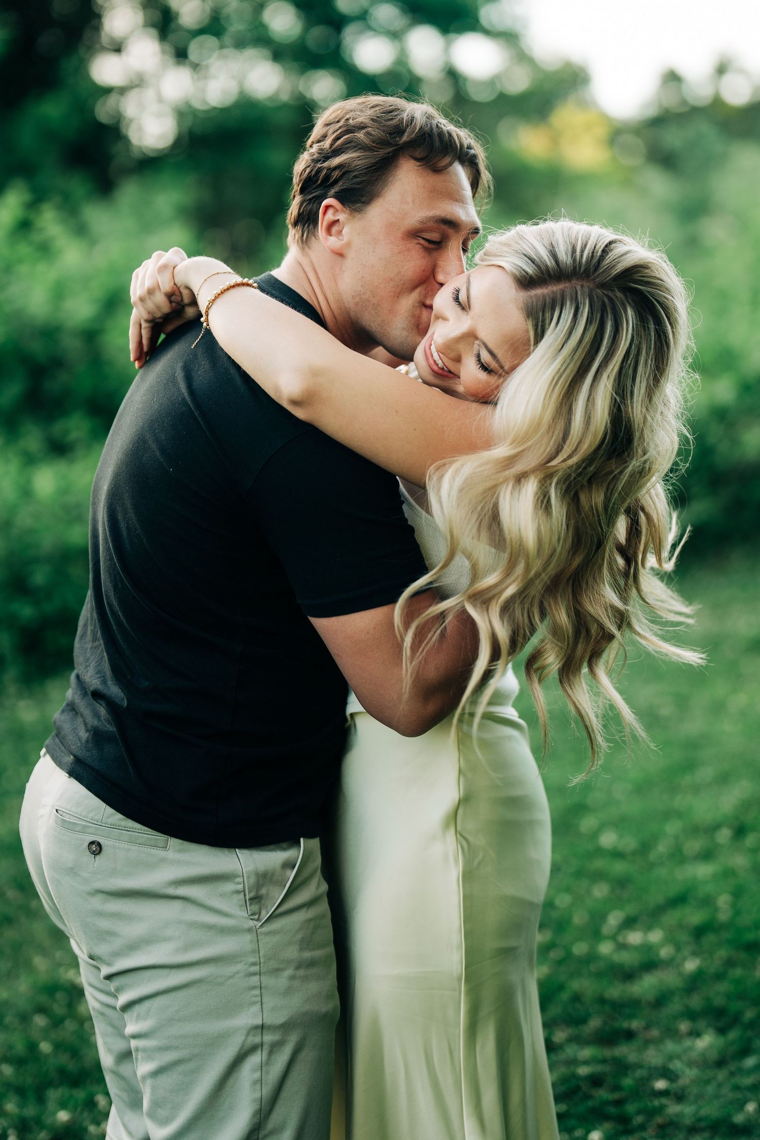 A couple embraces tenderly outdoors, the woman in a white dress leaning into a man in a black shirt amid lush greenery.