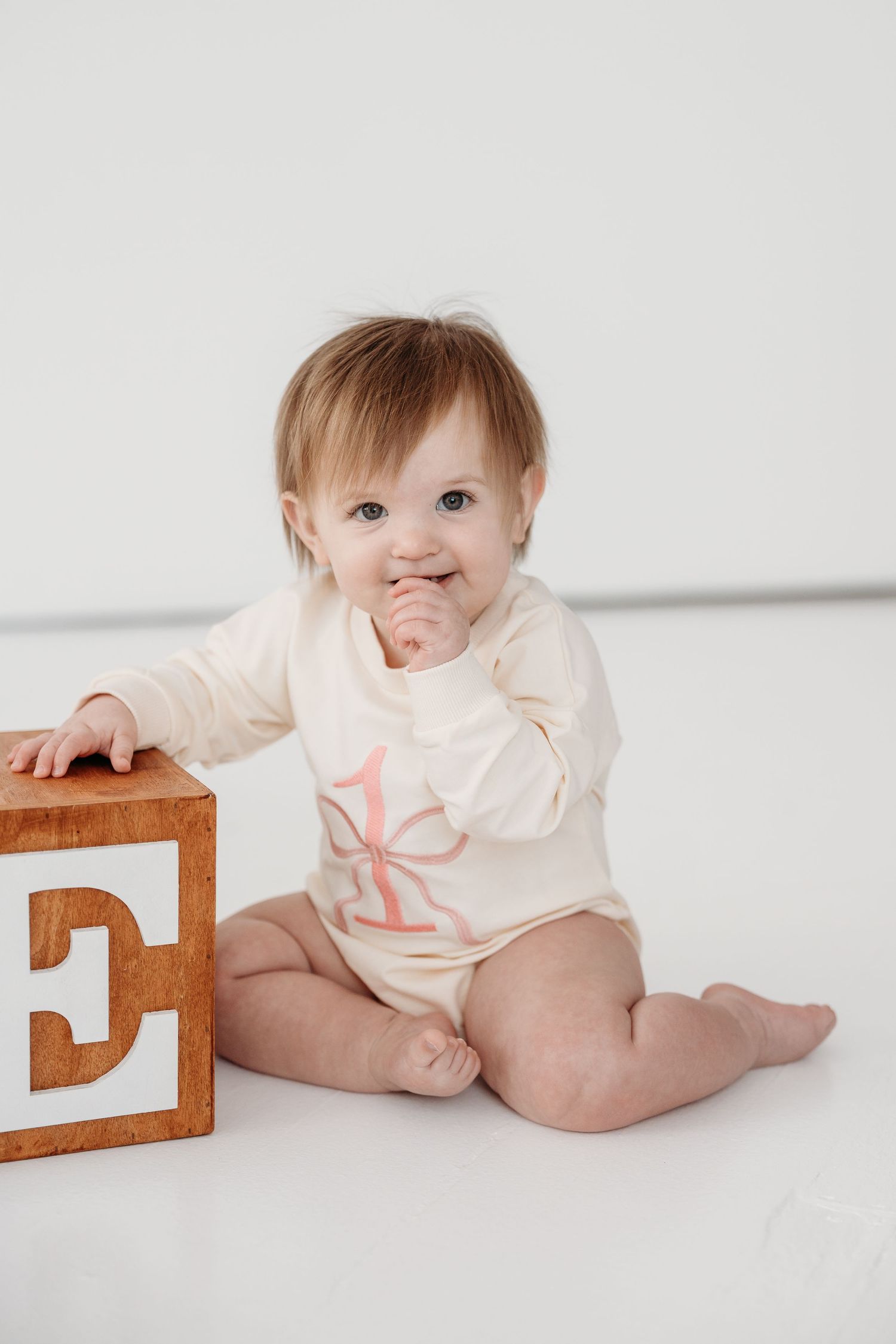Baby in cream-colored outfit sits on white background with wooden number blocks.