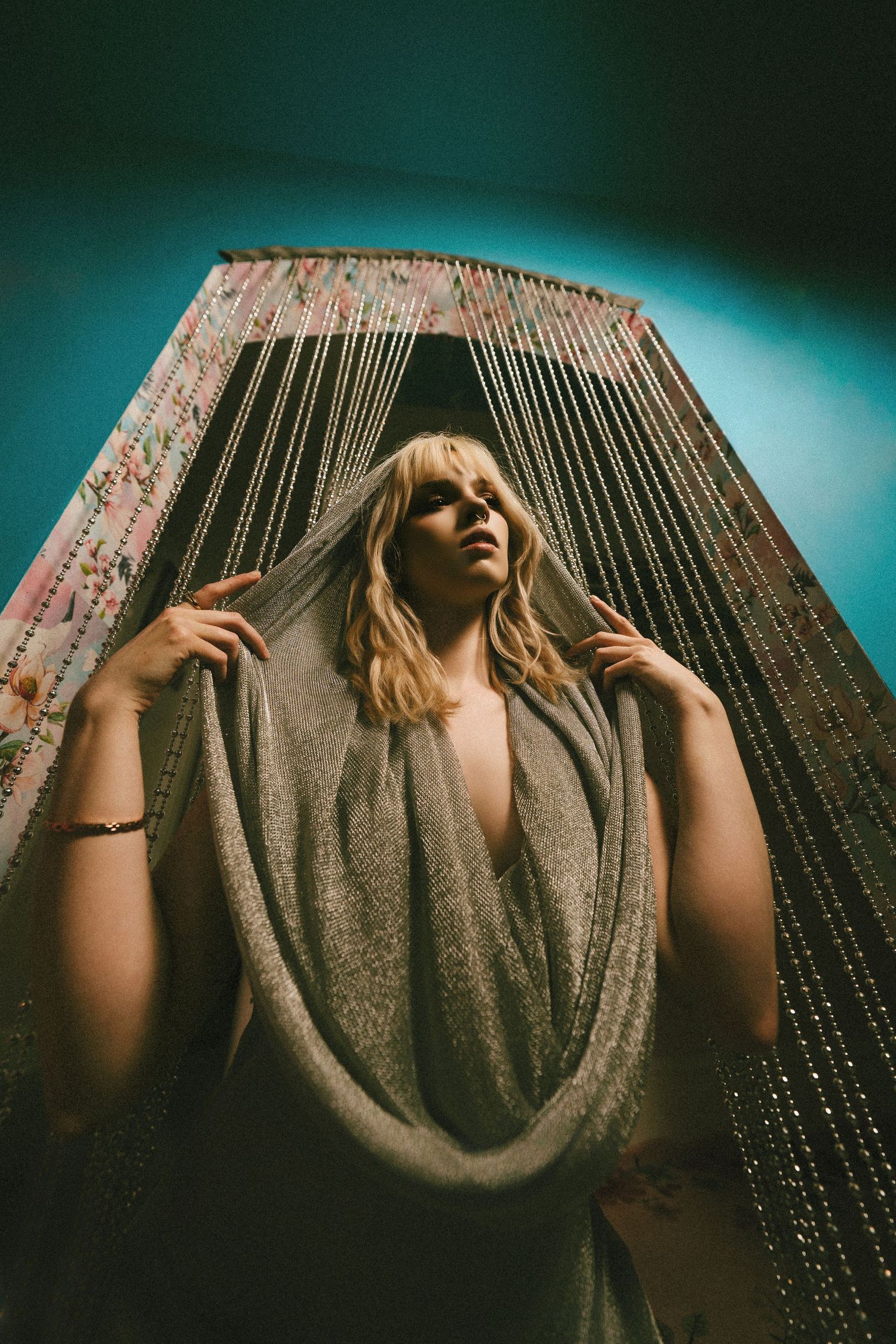Artistic portrait against beaded curtain backdrop with moody teal lighting and draped fabric creating dramatic atmosphere.