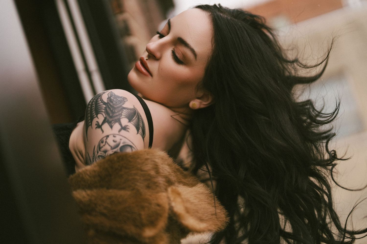 A person with flowing dark hair and tattoos poses with a brown cat in a warm-toned artistic portrait.