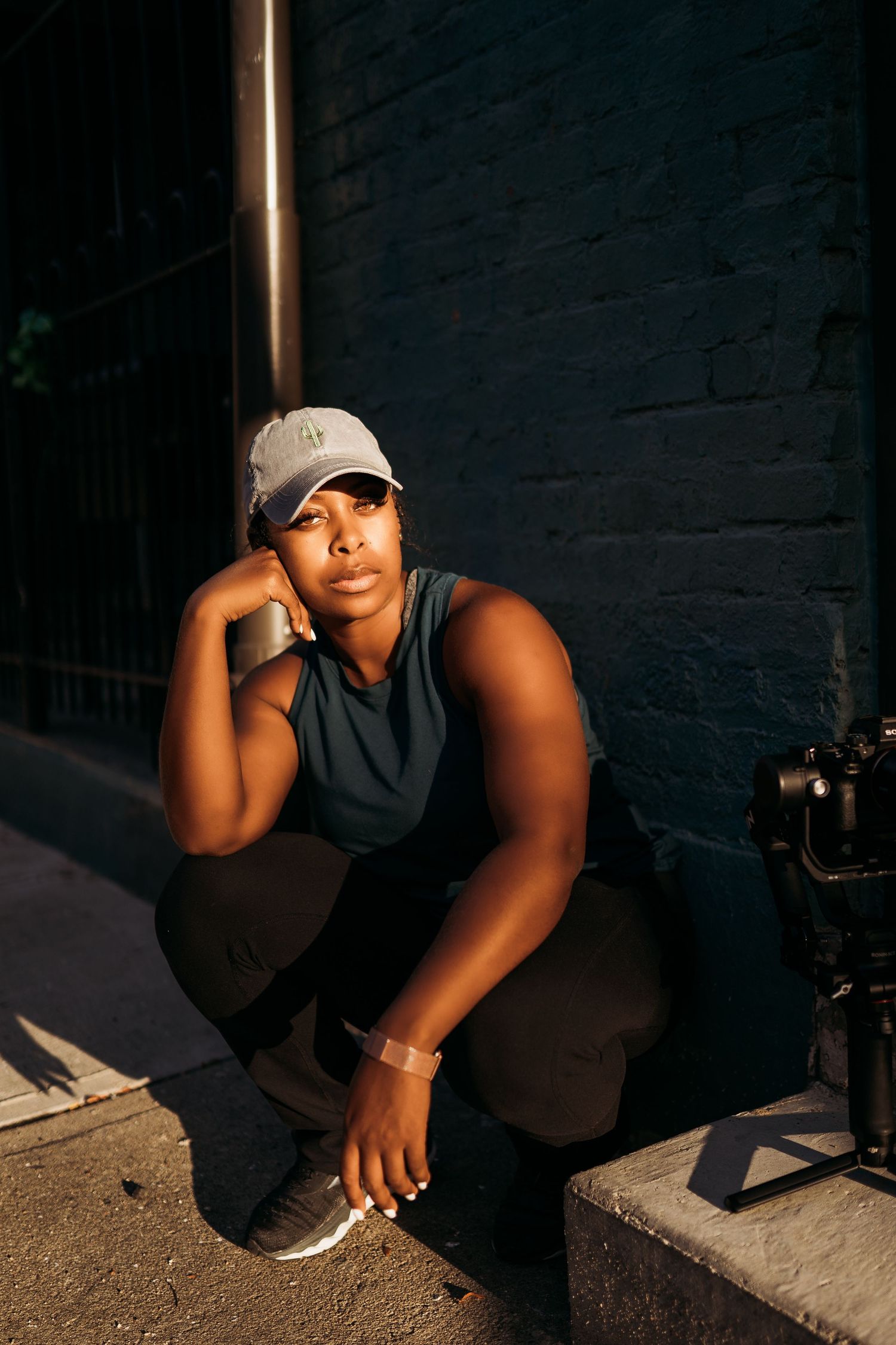 A person in a dark tank top and baseball cap sits in dramatic lighting against a shadowy brick wall background.