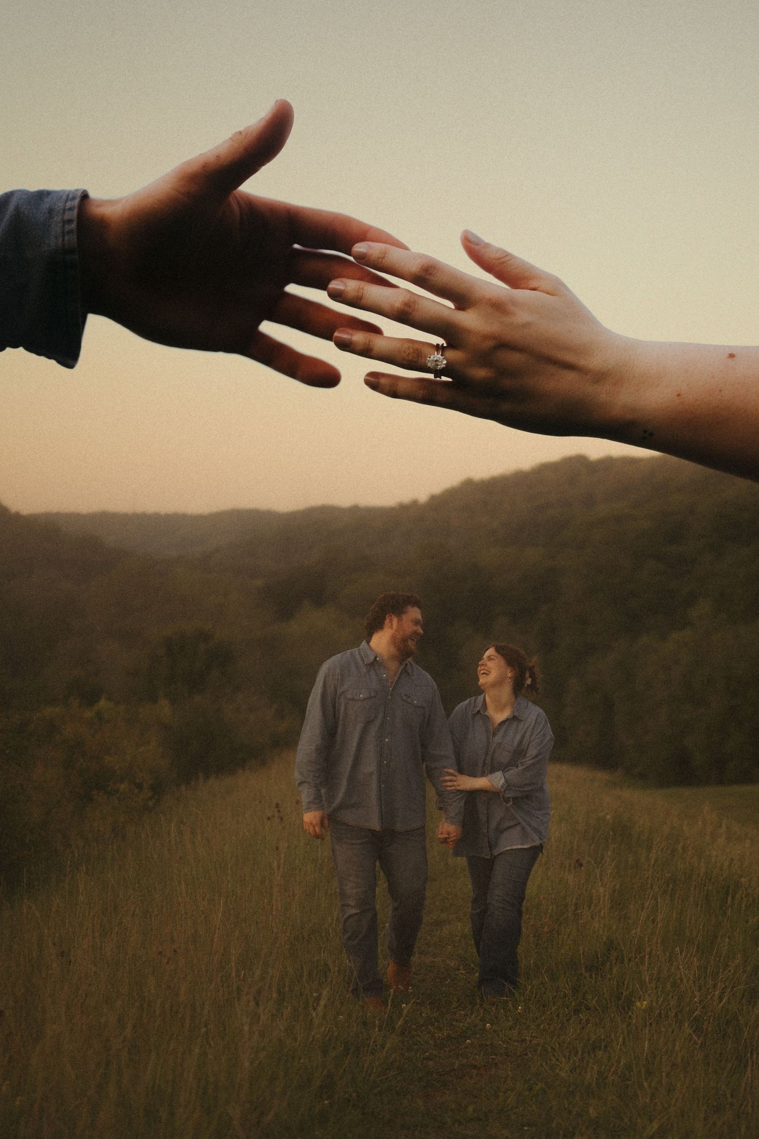 Couple walking in green meadow with hills at dusk, foreground shows hands touching with engagement ring visible.