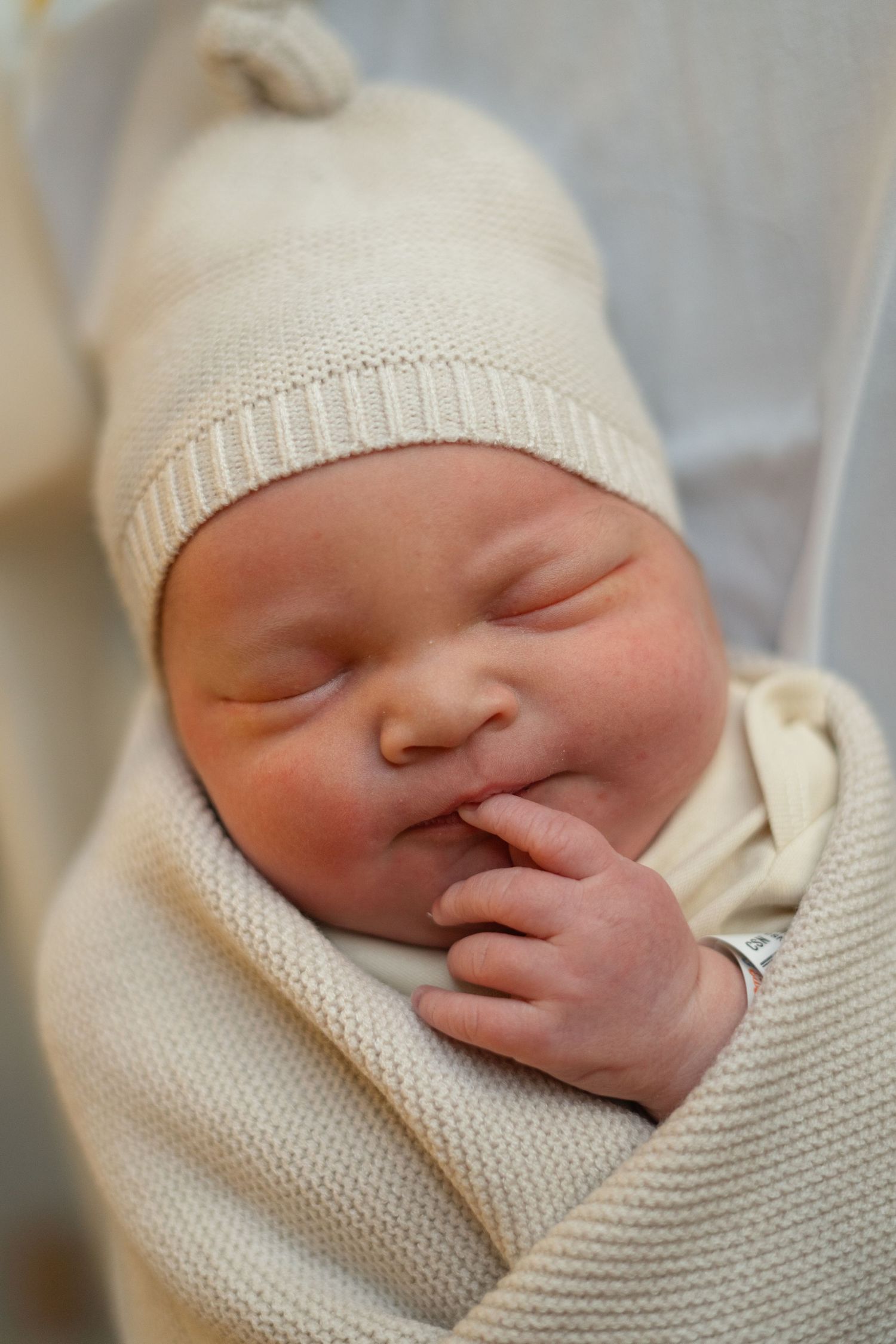 Sleeping newborn in cream knit beanie and sweater with hand near mouth makes peaceful expression.