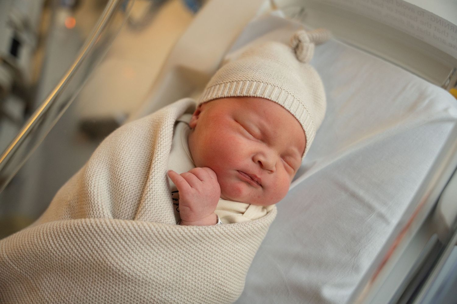 A sleeping newborn baby wrapped in a white blanket wearing a knit hat in a hospital bassinet.