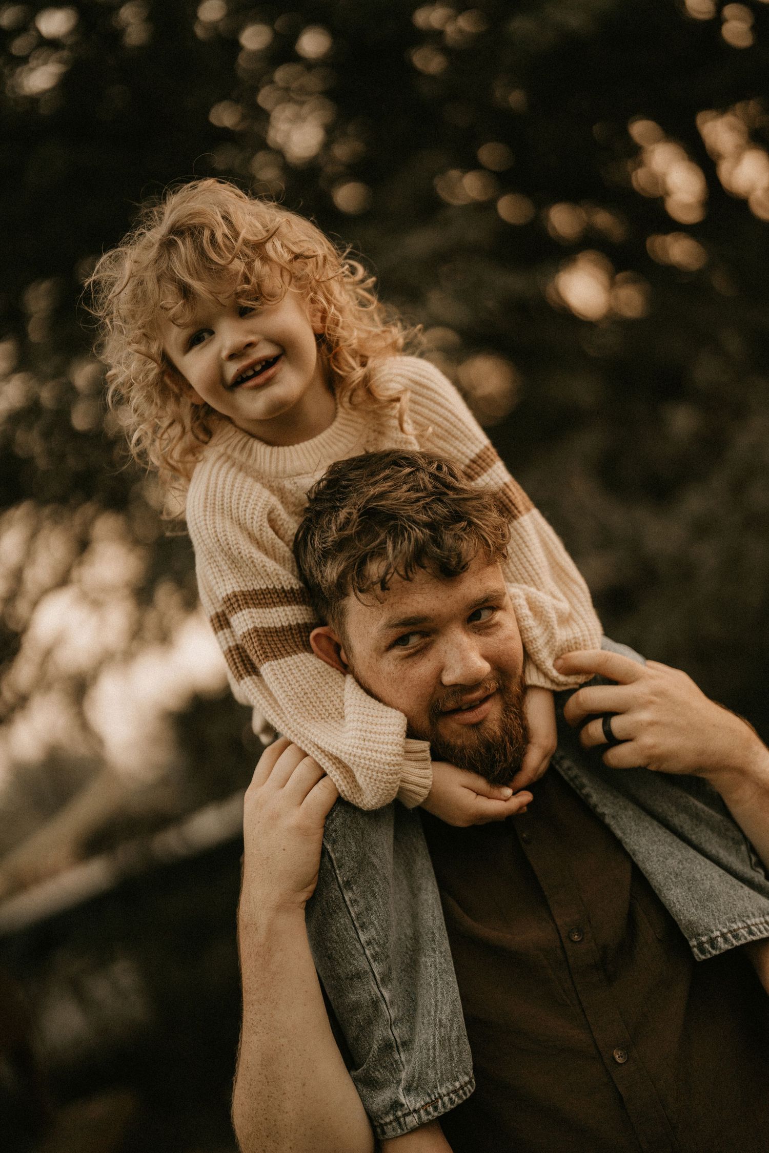 A bearded man smiles as a curly-haired child sits on his shoulders, both wearing sweaters outdoors.