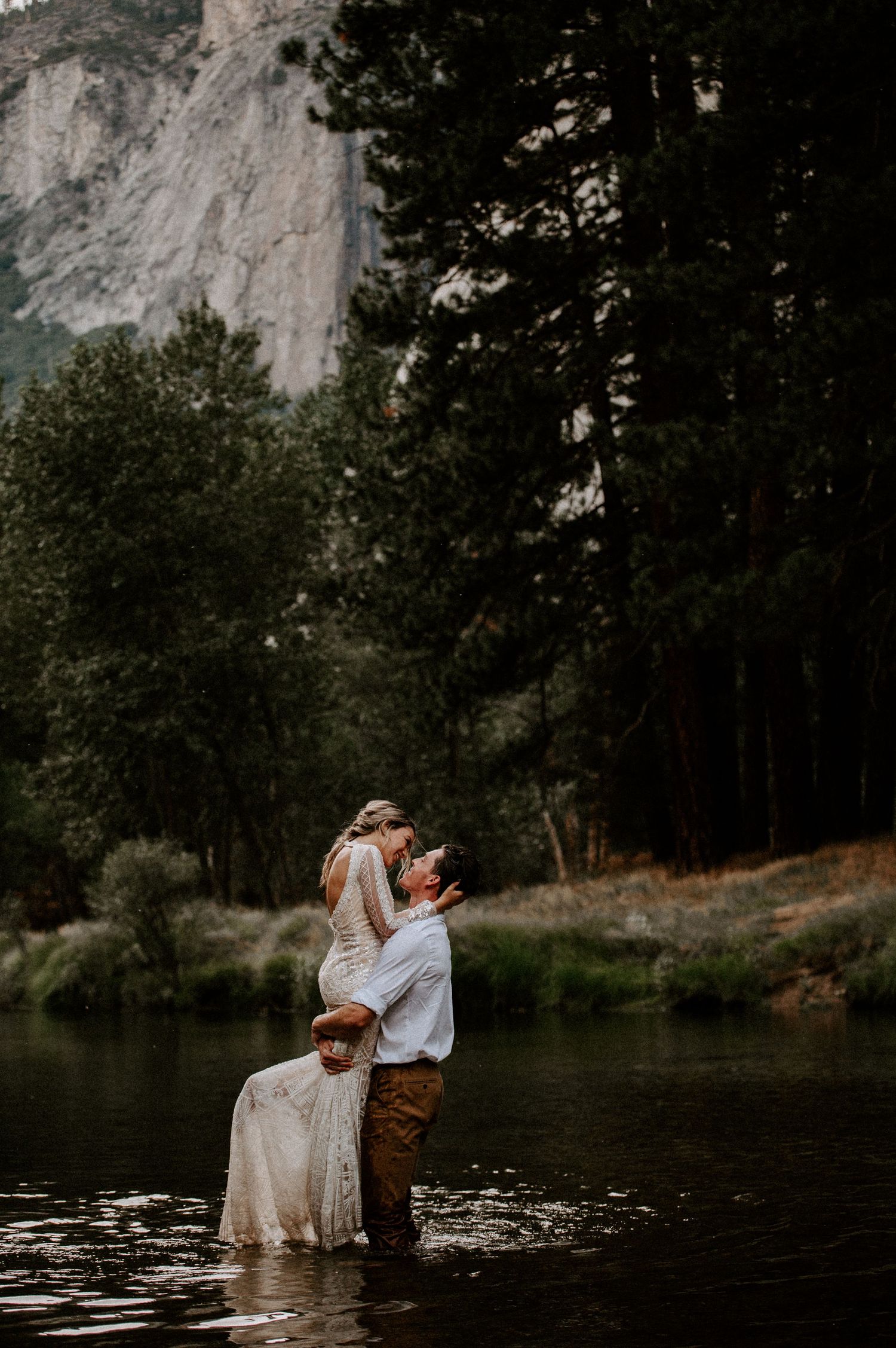 A romantic couple embraces while standing in a serene mountain lake surrounded by towering cliffs and forest at sunset.
