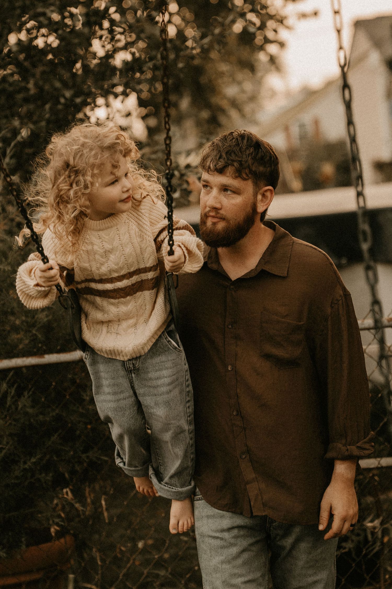 A bearded father watches his curly-haired toddler sitting on a backyard swing, both wearing casual autumn outfits.