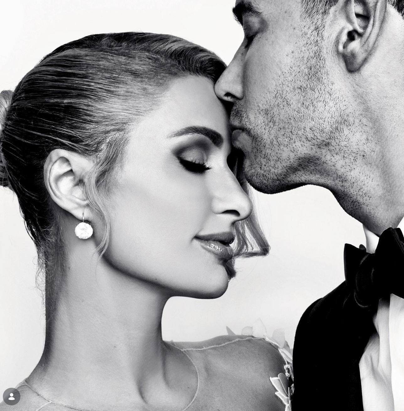 Black and white portrait of a groom in a tuxedo and bow tie kissing the forehead of a bride with closed eyes and pearl drop earrings.