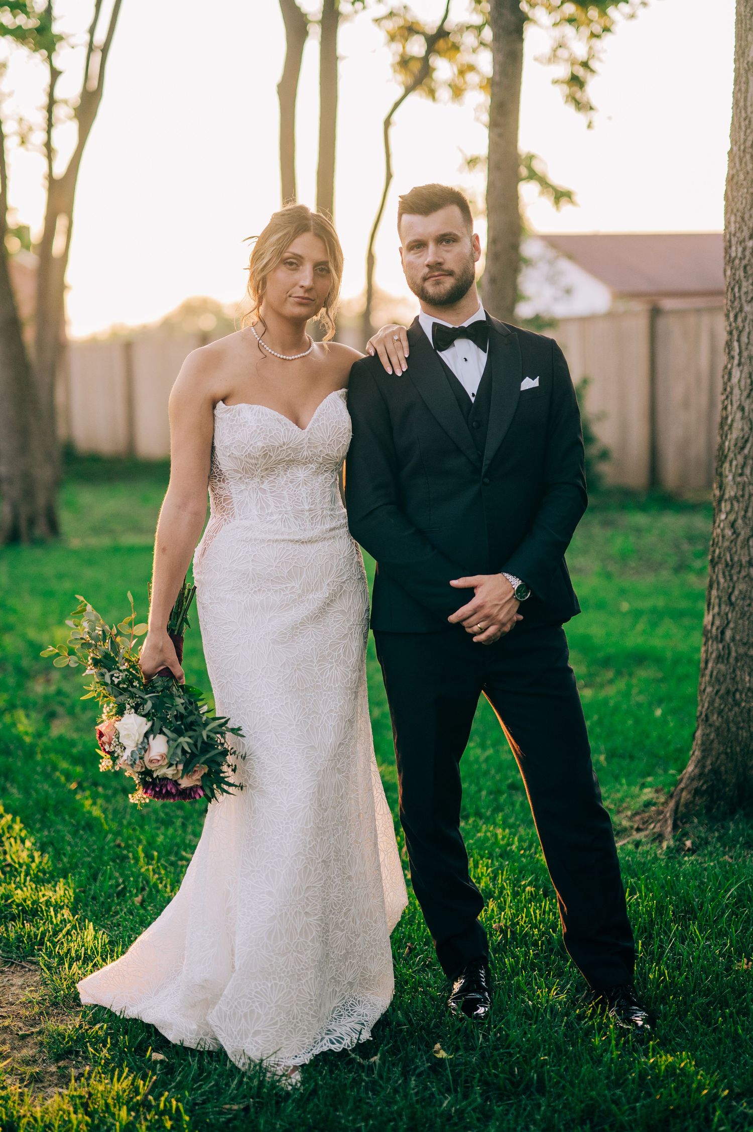 Bride in white lace gown holding bouquet stands with groom in black tuxedo outdoors at golden hour.