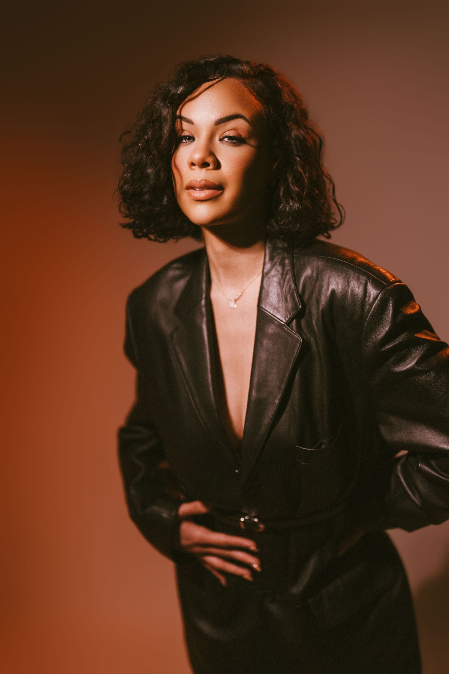 A person in a black leather blazer poses against a warm brown background in a studio photoshoot.
