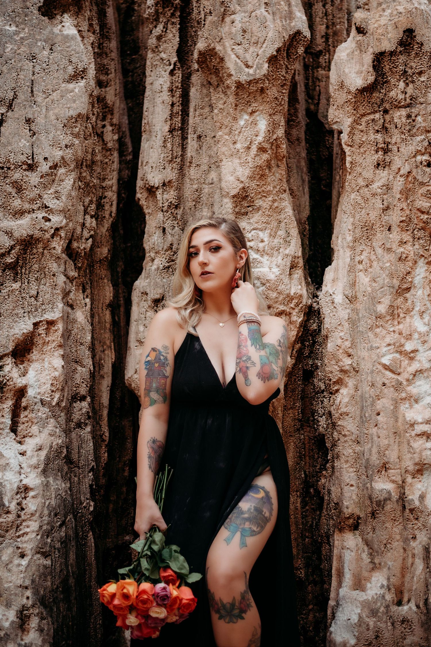 A woman in black poses against a rustic stone wall with tattoos visible on her arms and red flowers as props.