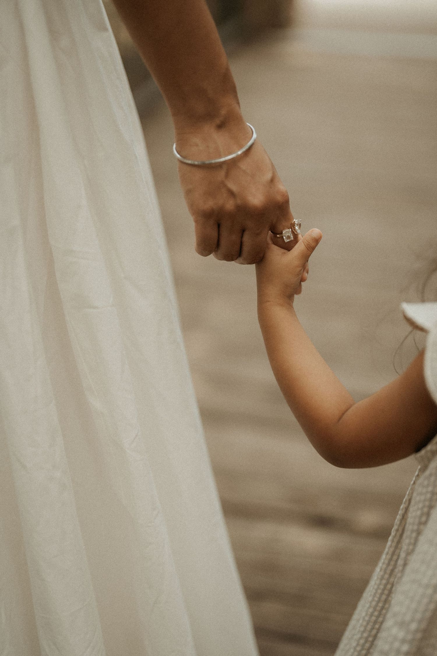 Adult hand with silver bracelet and ring gently holds a small child's hand, wearing white dress.