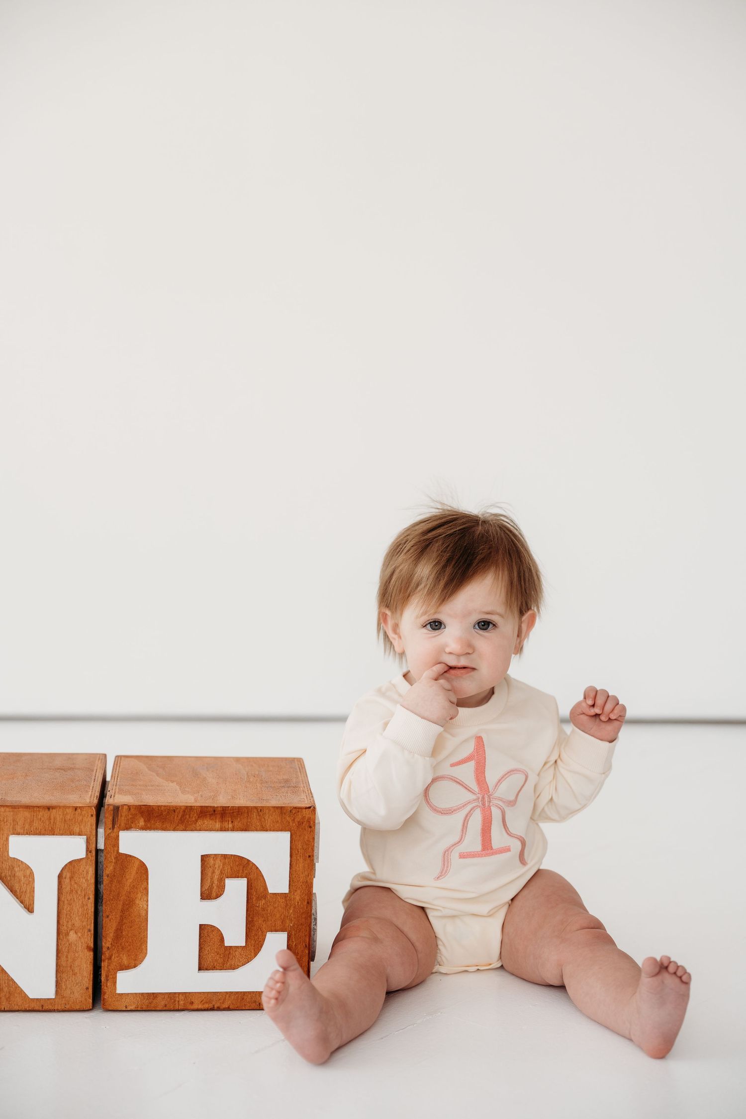 A small child in cream-colored clothing sits on a white floor next to wooden alphabet blocks spelling ONE.