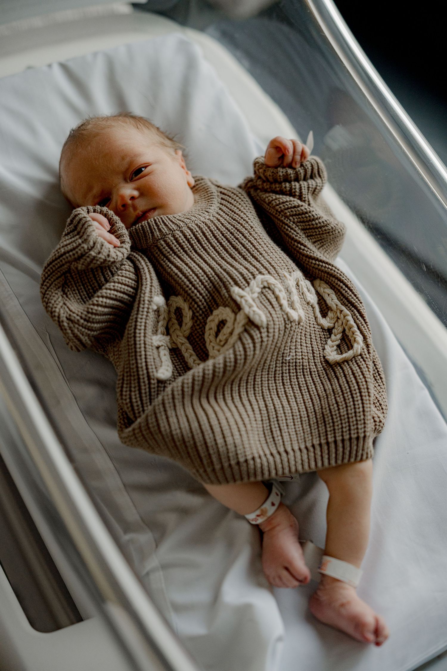 Baby wearing cozy knit brown sweater rests on white bedding in soft lighting.