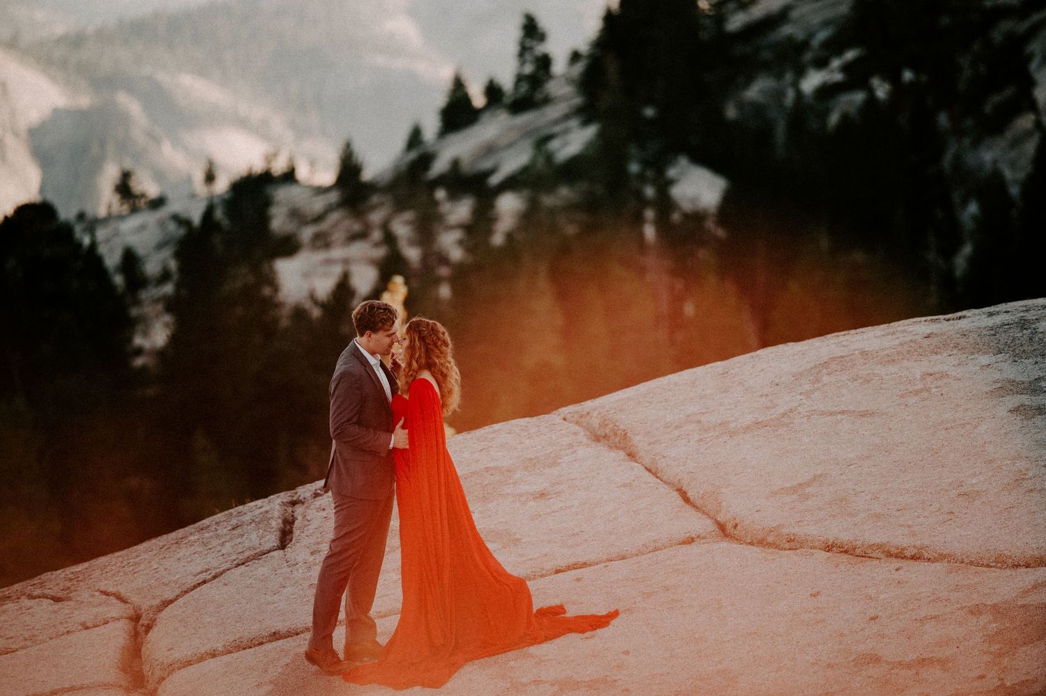 A couple in formal attire embraces on a snowy mountain ledge at sunset with pine trees in the background.