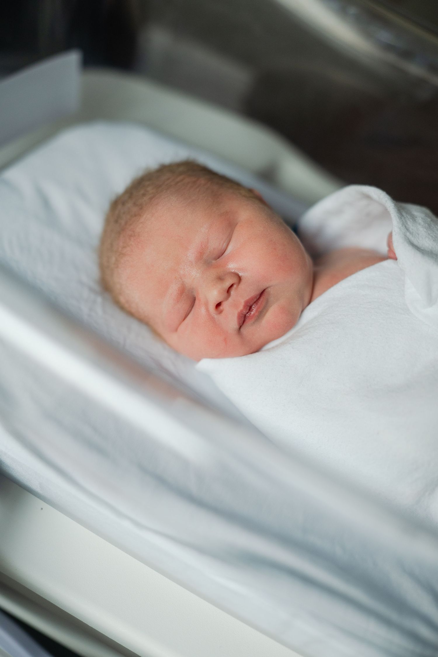 A peaceful sleeping newborn baby wrapped in soft white blankets in a hospital bassinet.