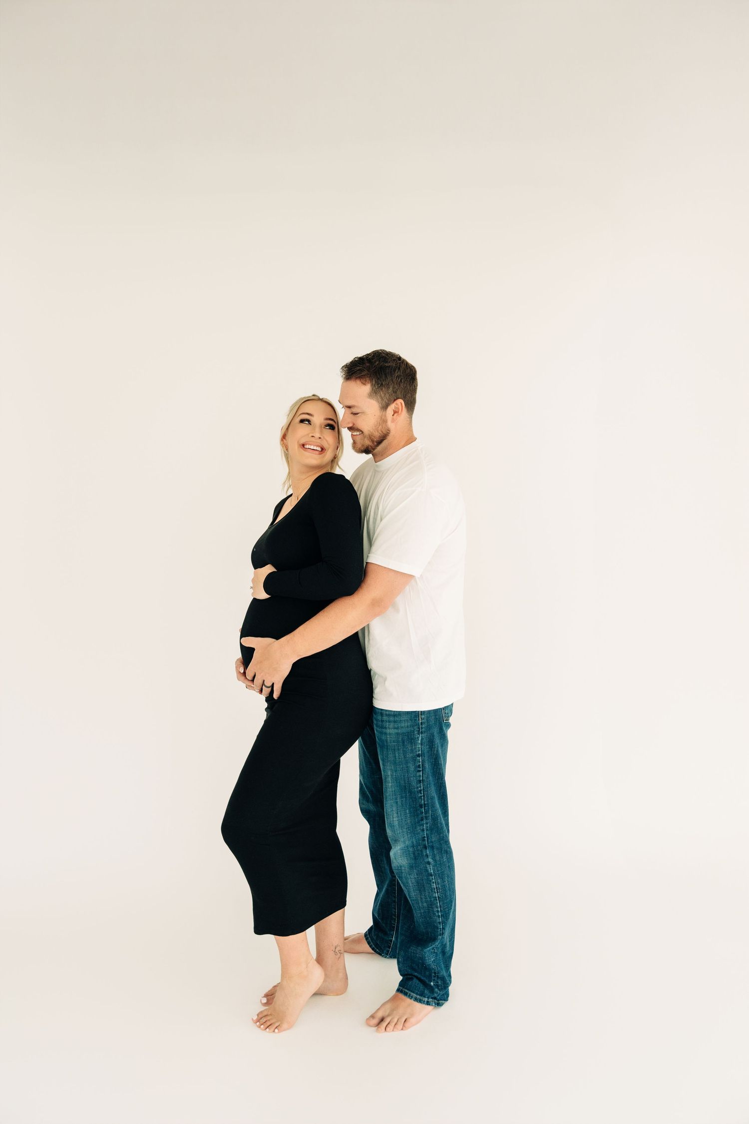A couple embraces in a minimalist white studio setting, wearing casual attire and standing barefoot together.