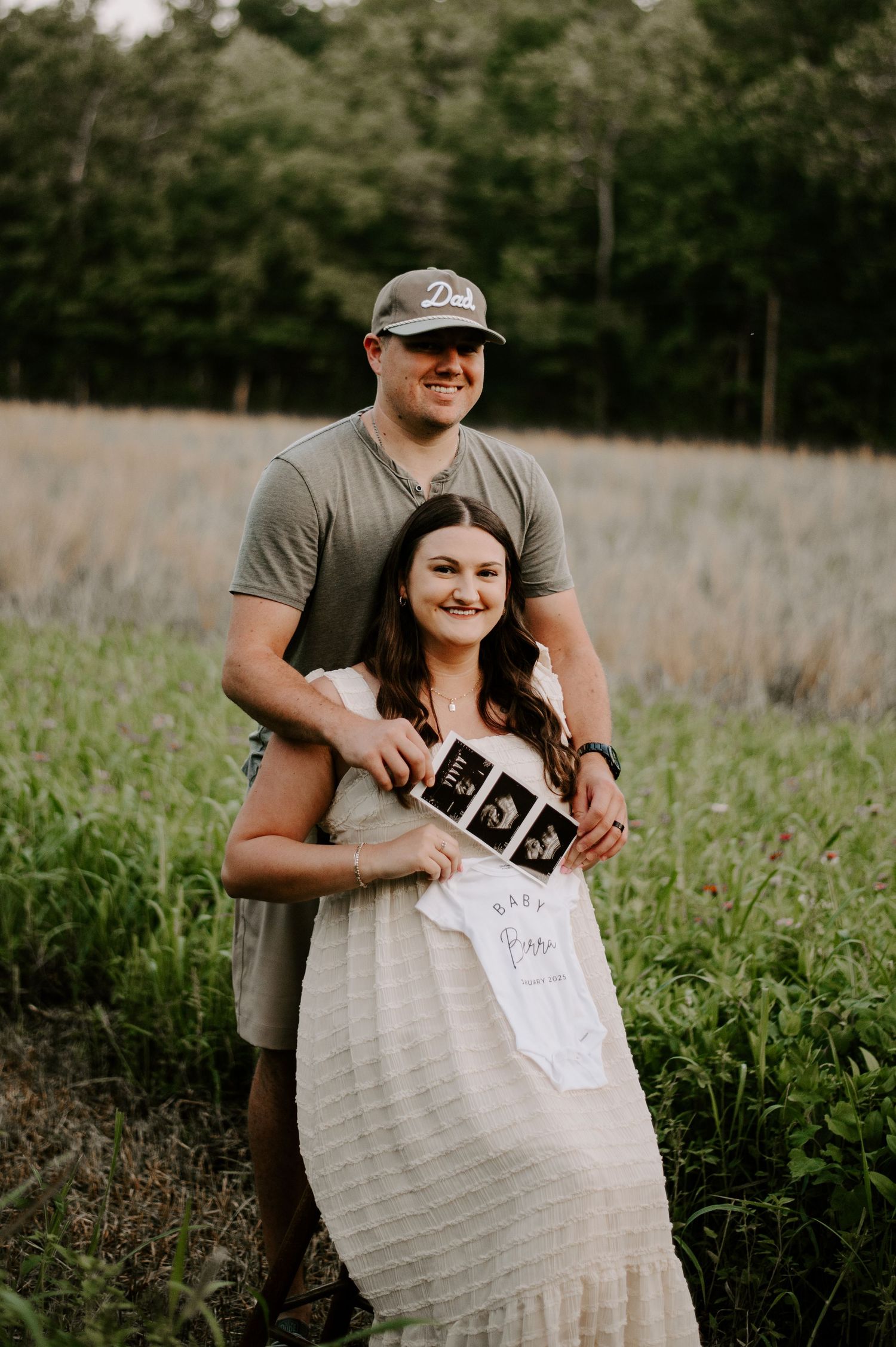 A couple stands in a grassy field holding ultrasound photos during a pregnancy announcement photoshoot.