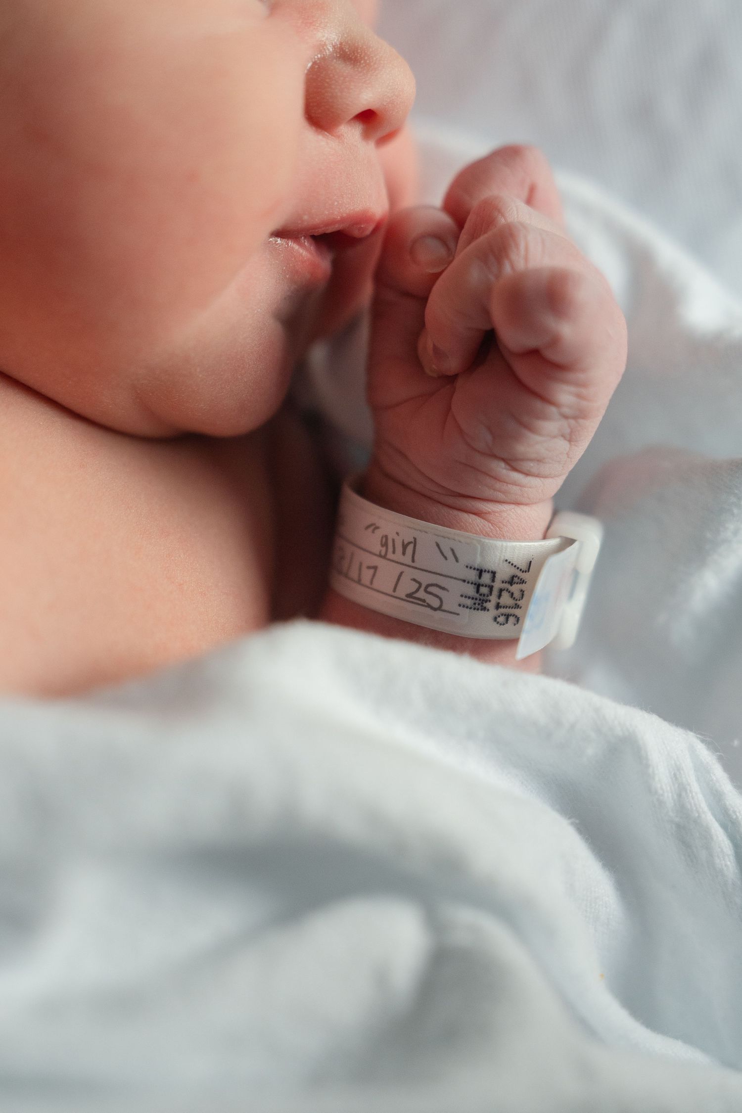 Close-up of newborn baby's hand with hospital ID bracelet sucking thumb while resting on white blanket.