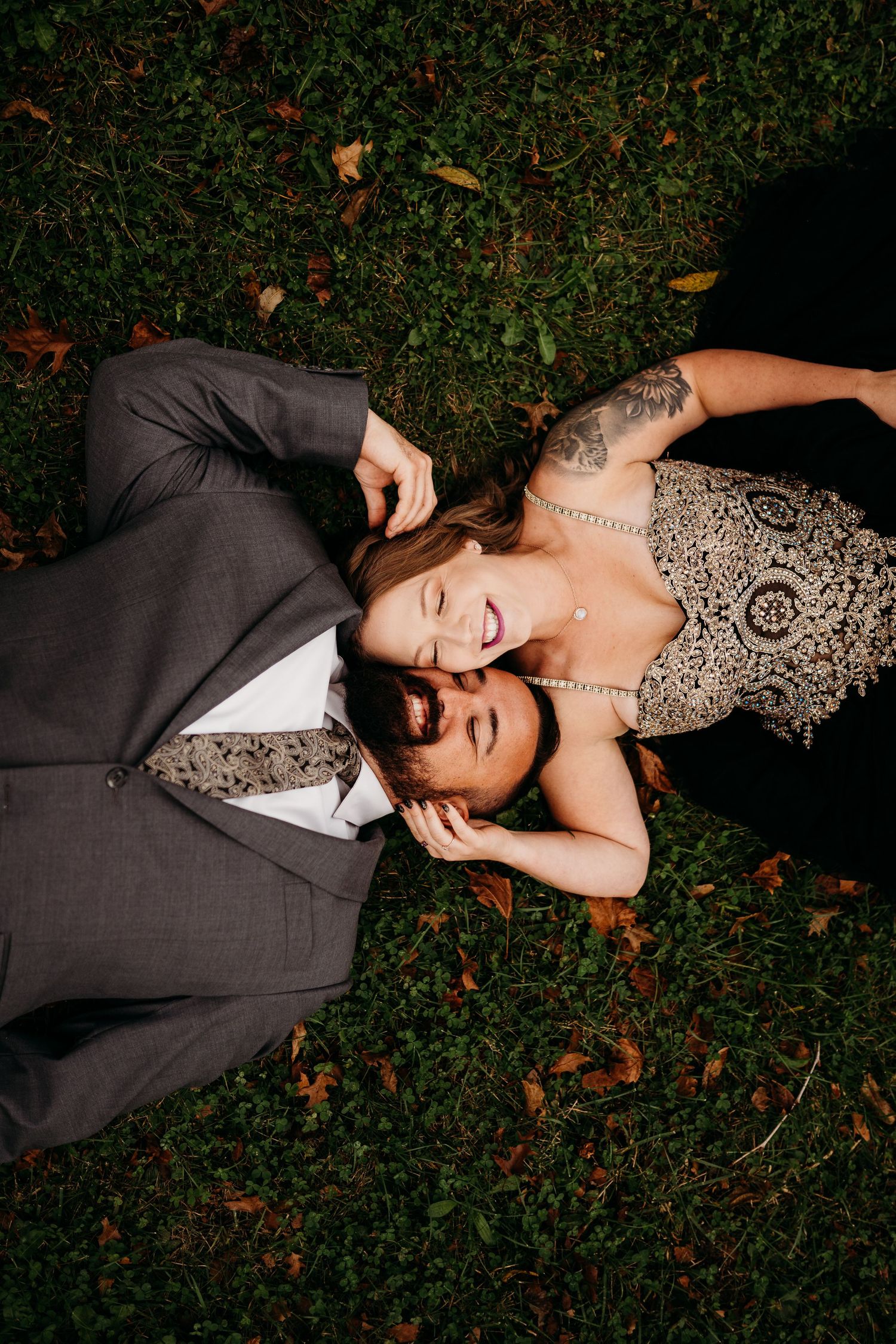A couple in formal attire lies on grass, with the woman in a lace dress resting her head on the man's chest.