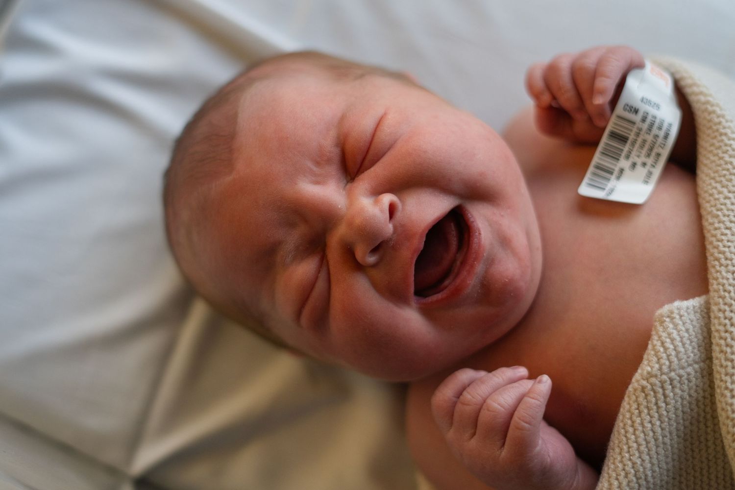 A crying newborn baby with clenched fists lies on white bedding in a hospital setting.