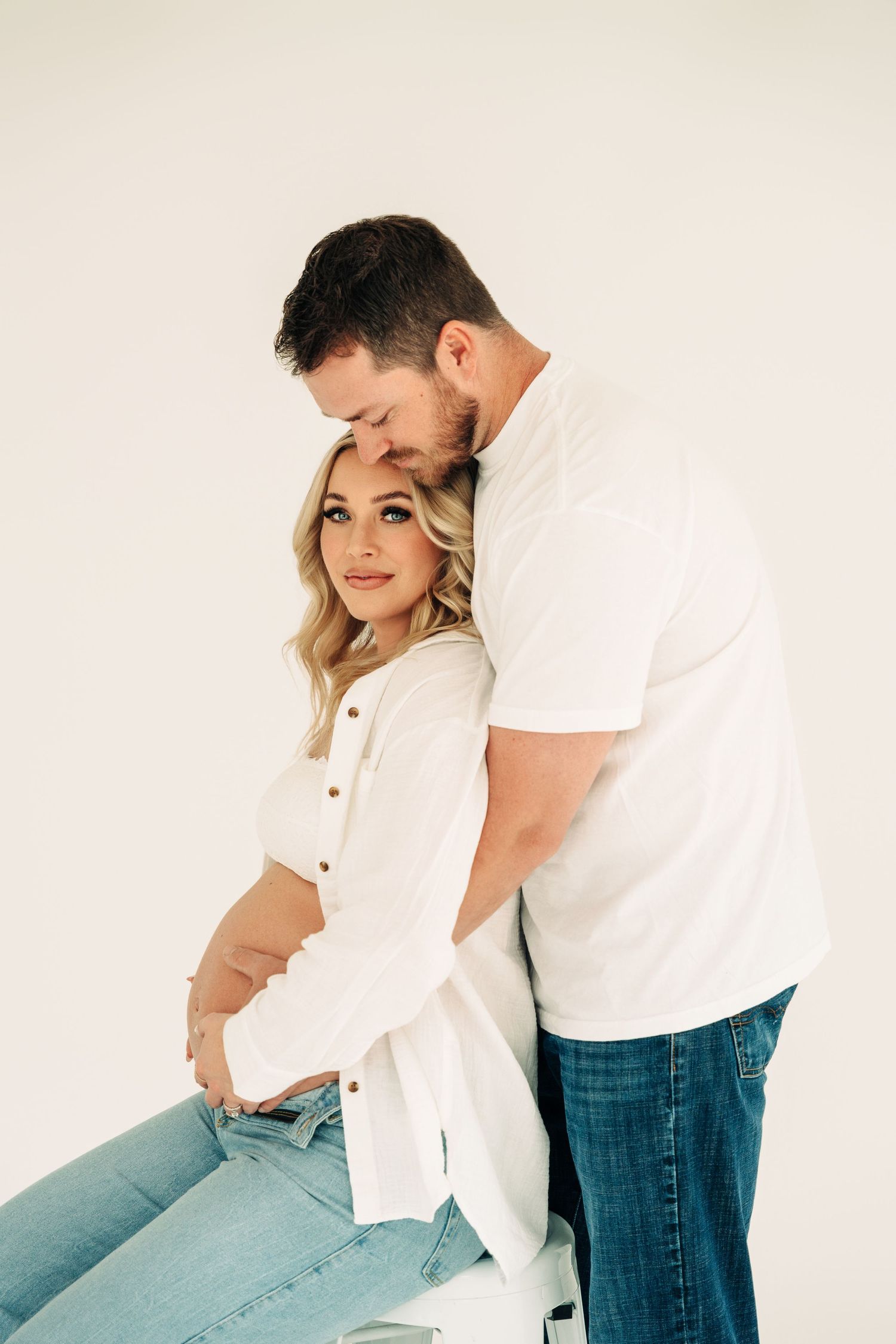 A couple in white shirts and jeans embrace in a minimalist studio setting against a light background.