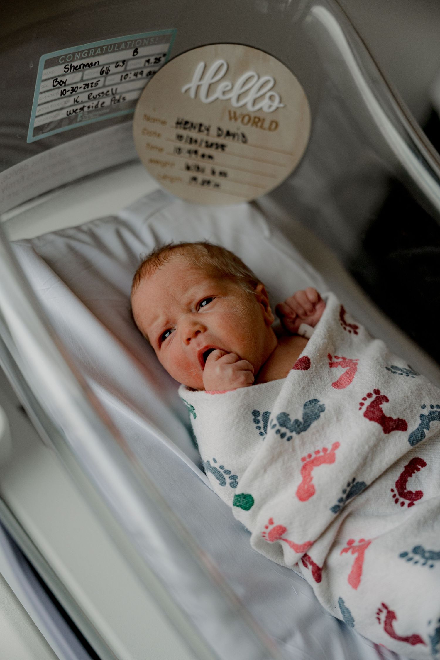 Newborn baby wearing white sleepwear with colorful dinosaur prints lies in hospital bassinet.