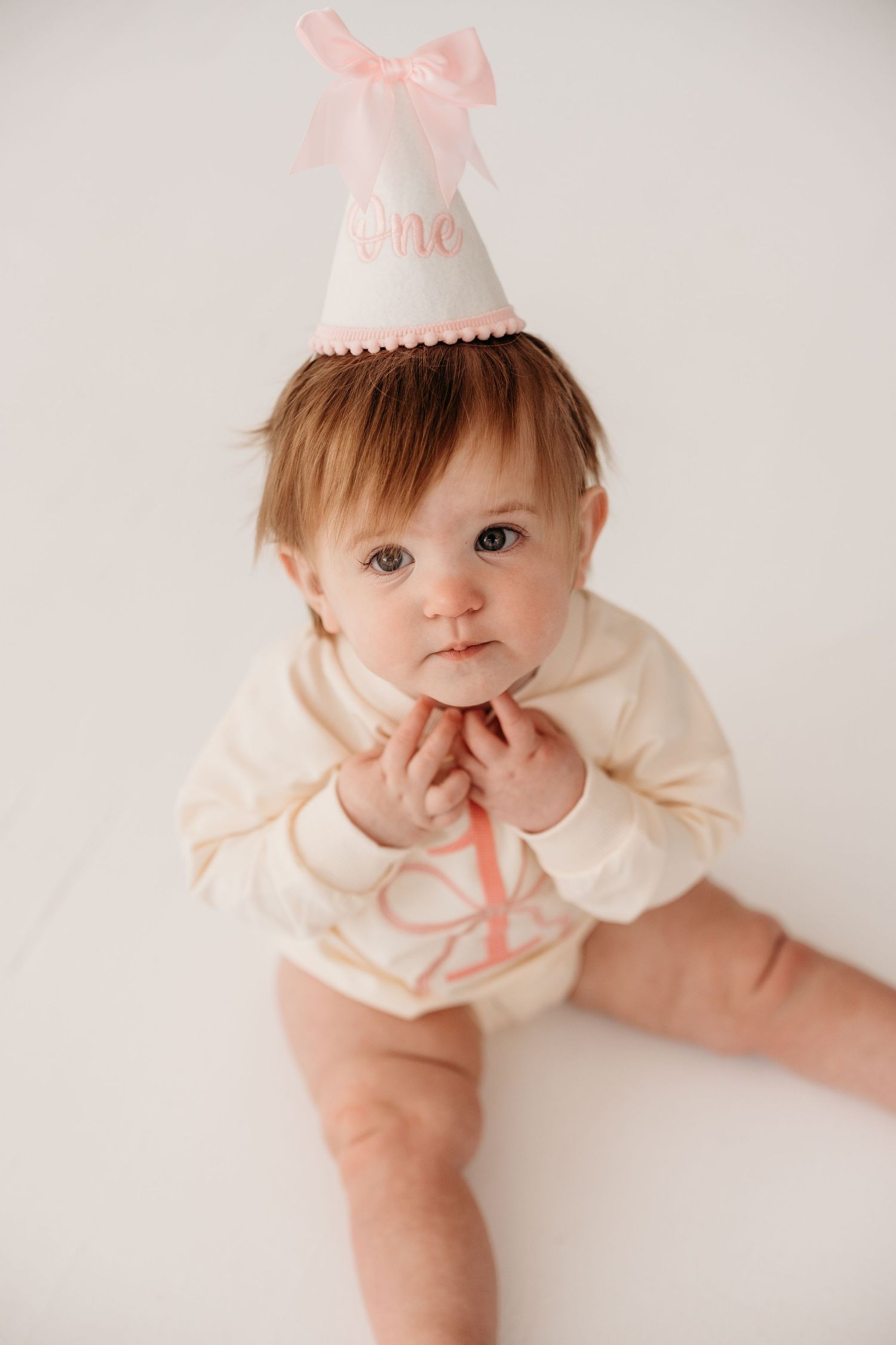 A baby in a cream-colored dress and party hat poses against a white background in a series of sweet portraits.