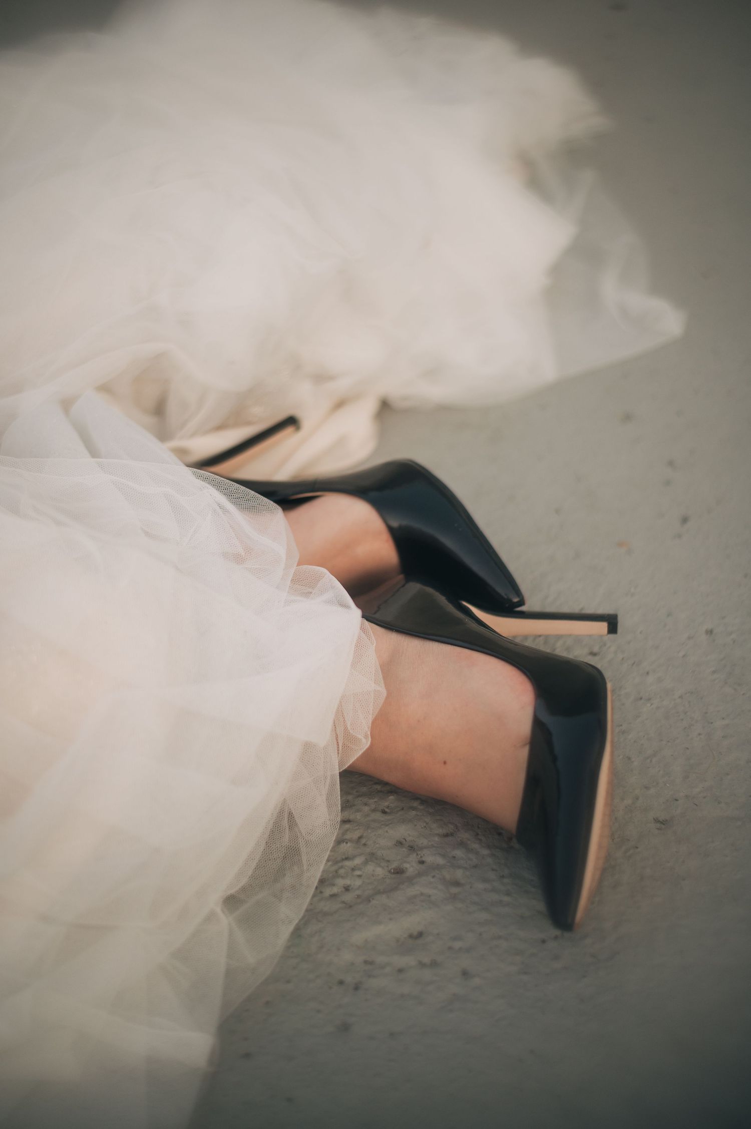 A woman's feet in black stiletto heels peek out from beneath flowing white tulle fabric of a wedding dress.