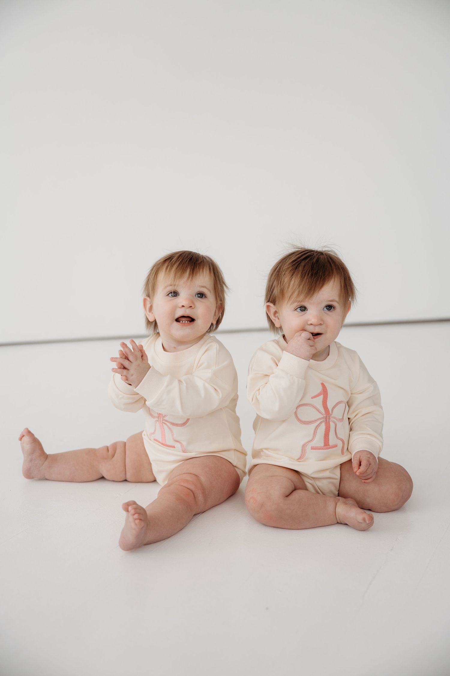 Two babies in matching cream colored outfits sit together on a white background in a series of sweet poses.