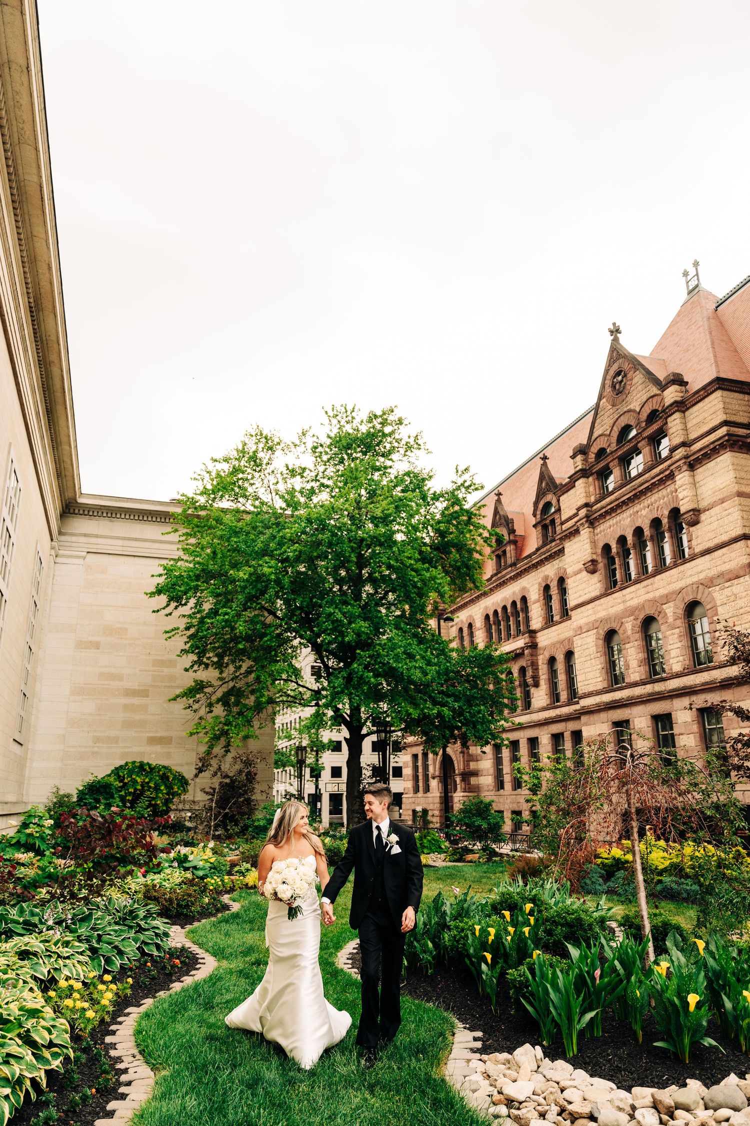 A panoramic view of a wedding couple walking along a stone path beside a historic Gothic-style building with lush gardens.