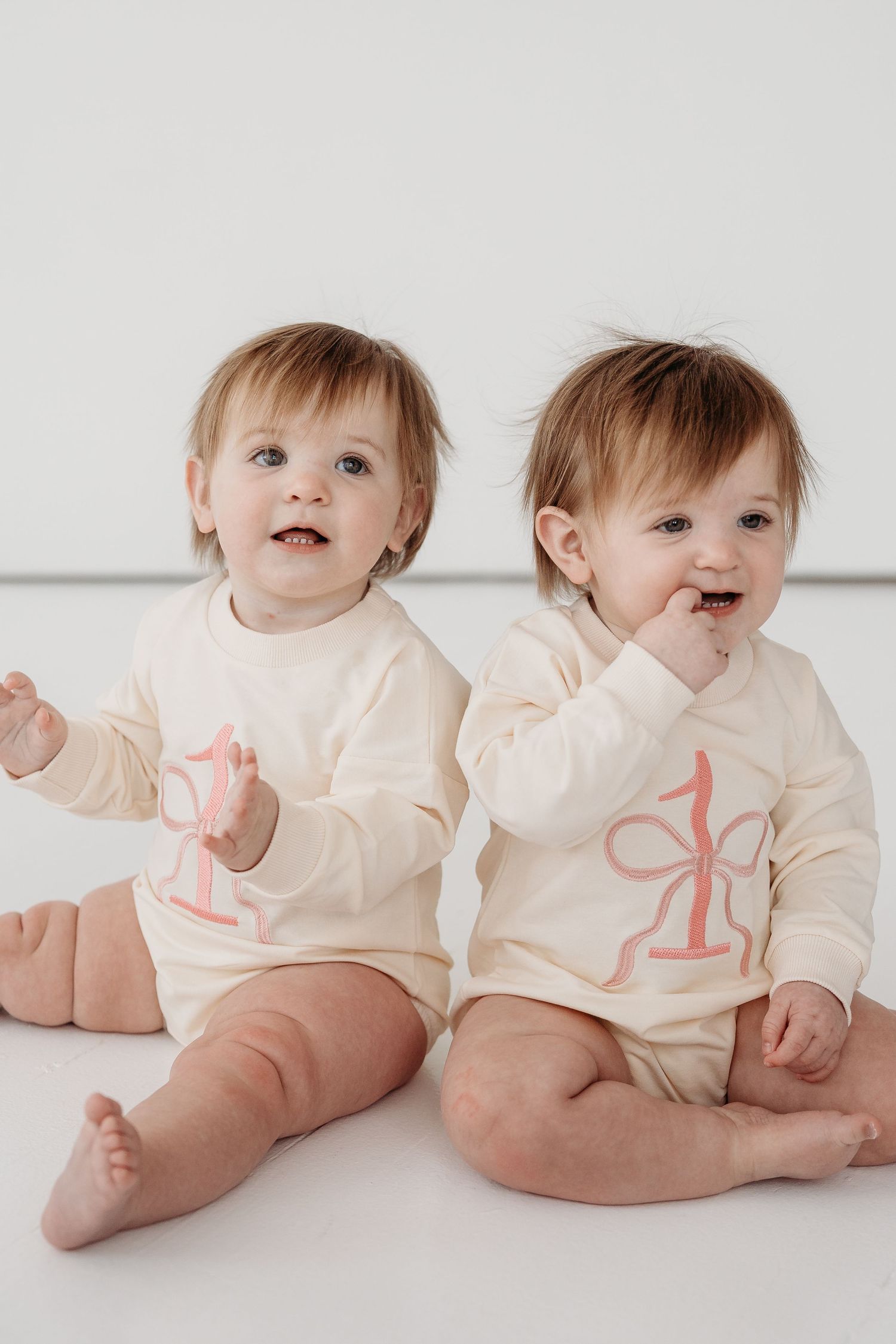 Two babies wearing cream-colored onesies with pink number one designs sit together on white background.