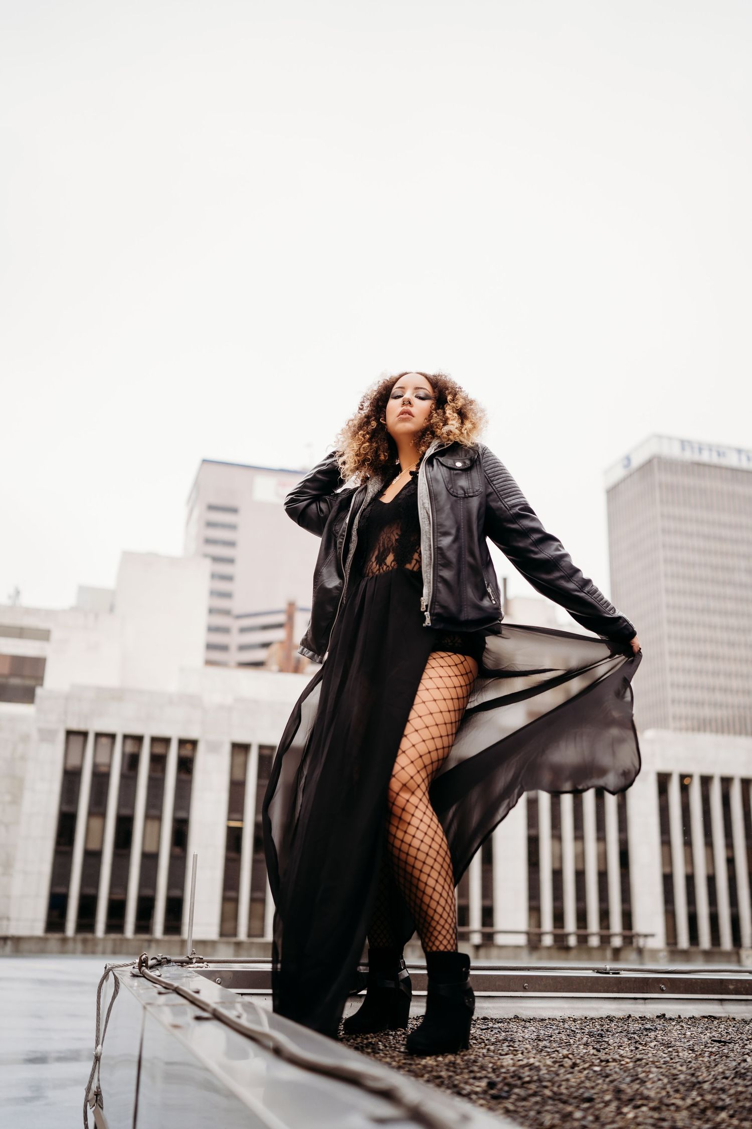 Woman in black leather jacket and fishnet stockings poses dramatically on urban rooftop with city buildings in background.