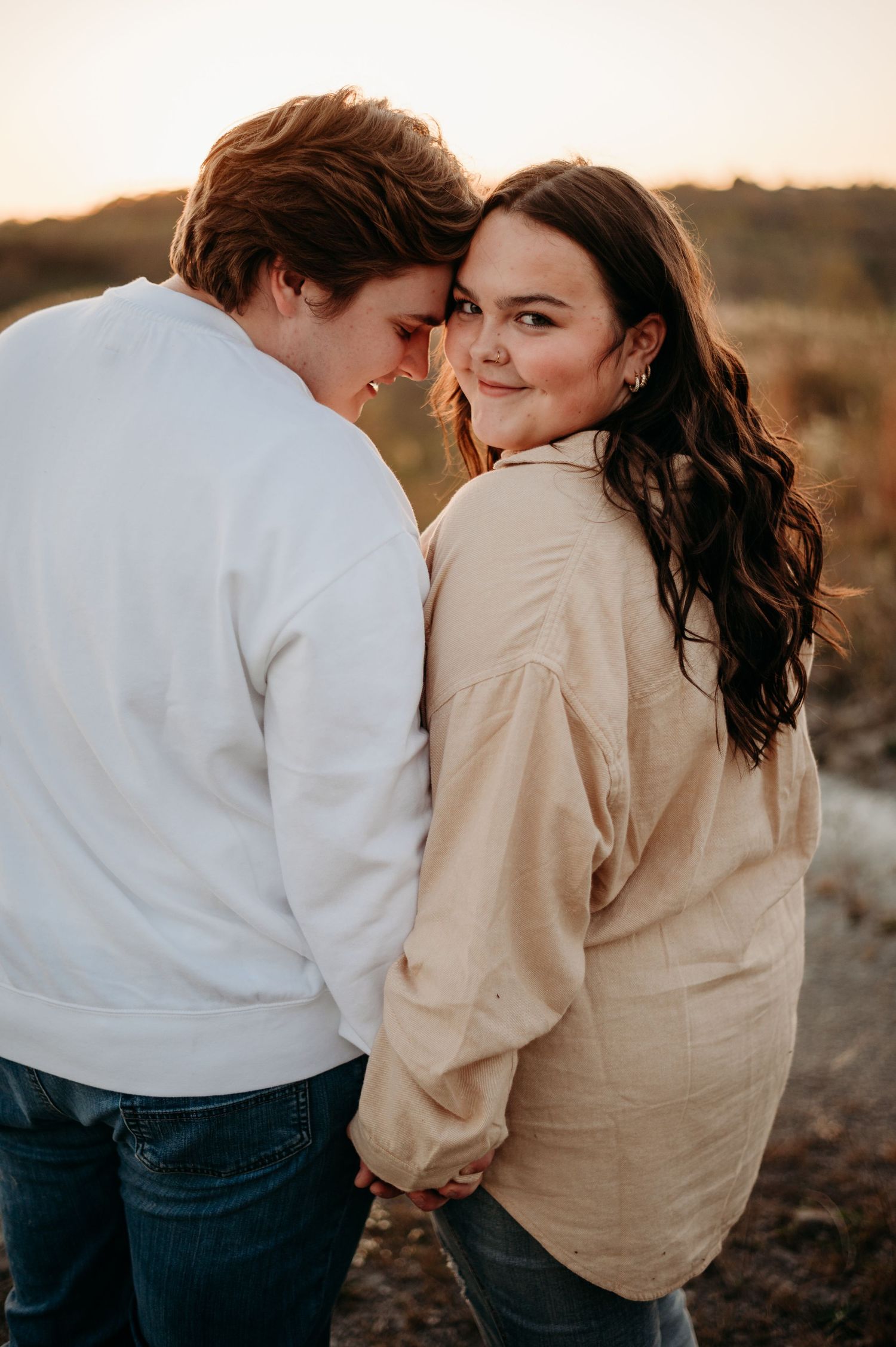 A couple embraces intimately at sunset, with the woman smiling over her shoulder in a warm, romantic outdoor setting.