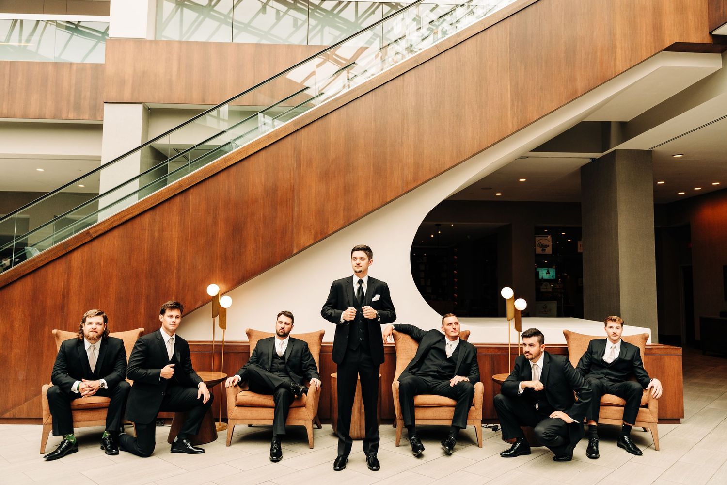 Wedding party in formal black suits poses elegantly in modern lobby with wood paneling and glass staircase.