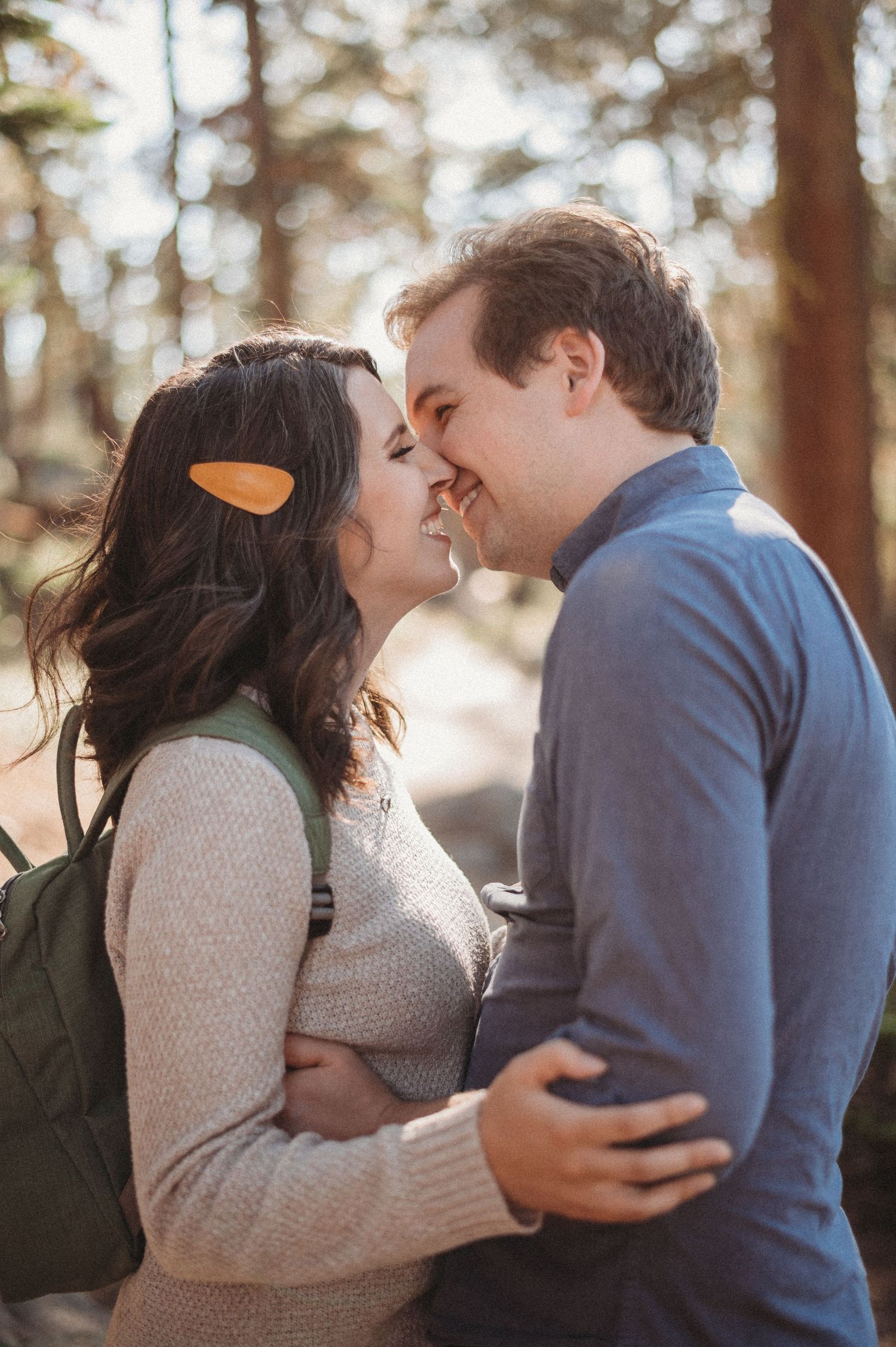 A couple embraces in a sunlit forest, the woman wearing a hair clip and the man in a blue sweater.