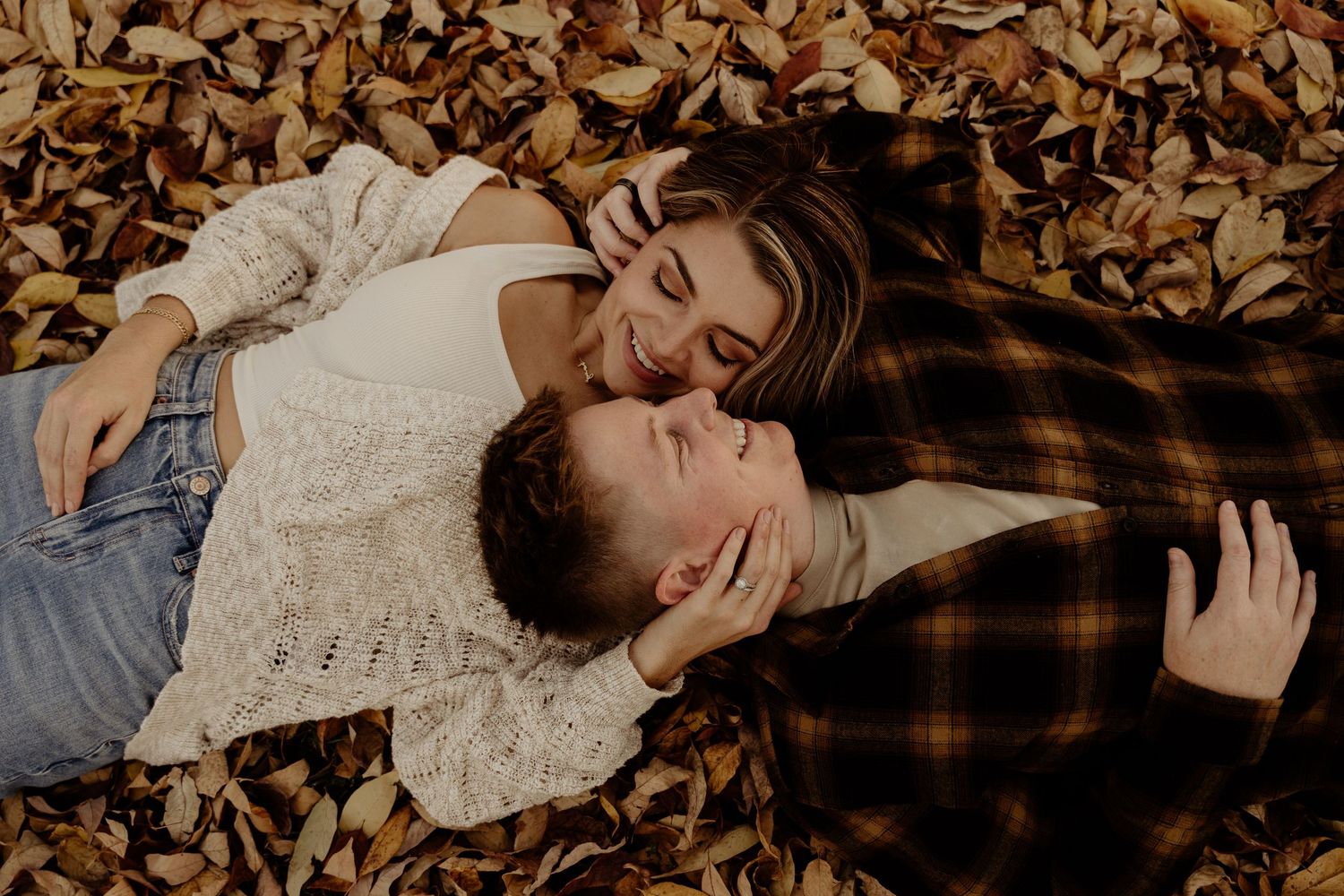 A couple lies intimately on a plaid blanket surrounded by autumn leaves in a romantic outdoor setting.