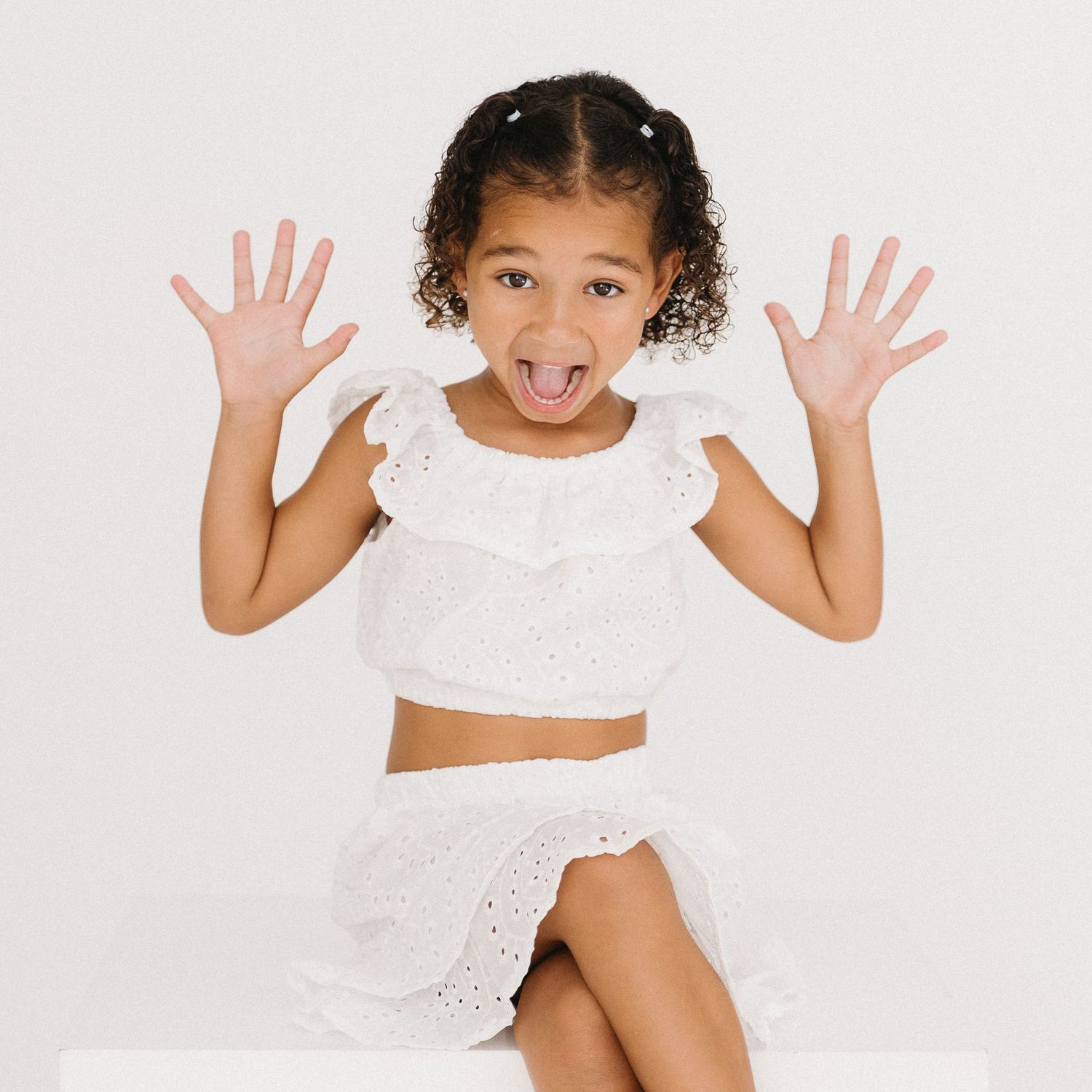 Energetic child in white outfit sitting and posing with raised hands against white background.