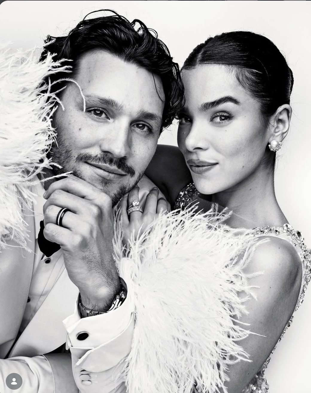 Black and white portrait of a smiling bearded man and a woman with slicked-back hair, posing close in glamorous white feathered attire.