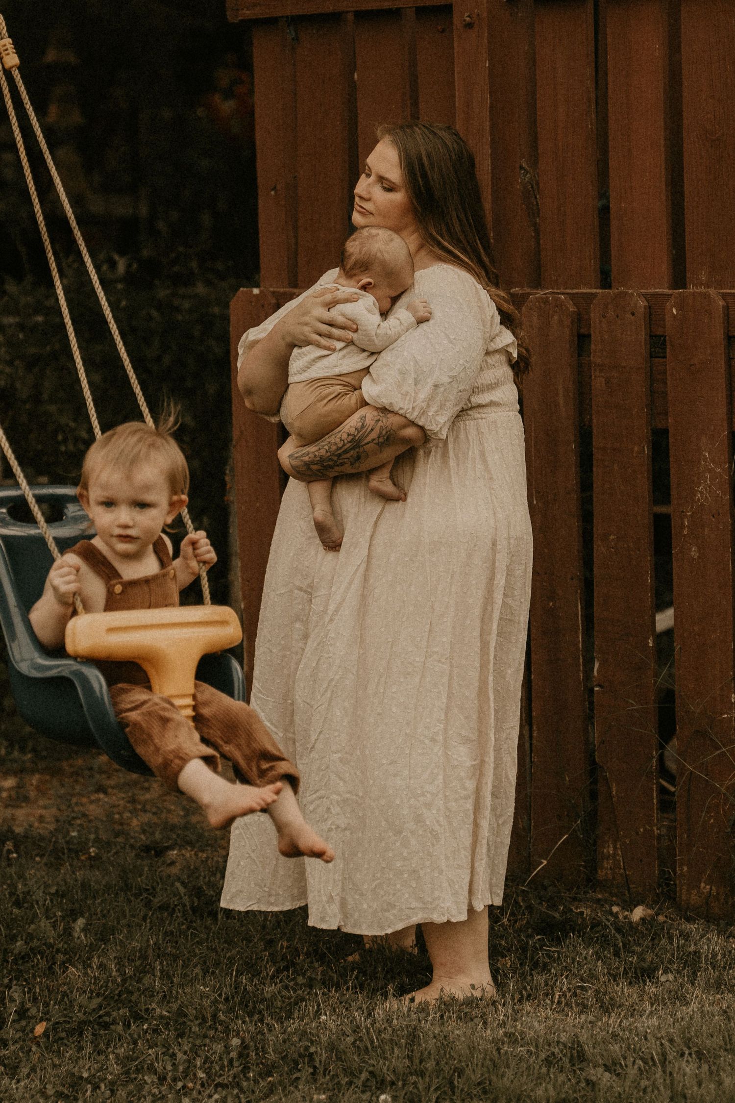 A mother in a cream dress holds a newborn while a toddler sits in a swing in a backyard setting.