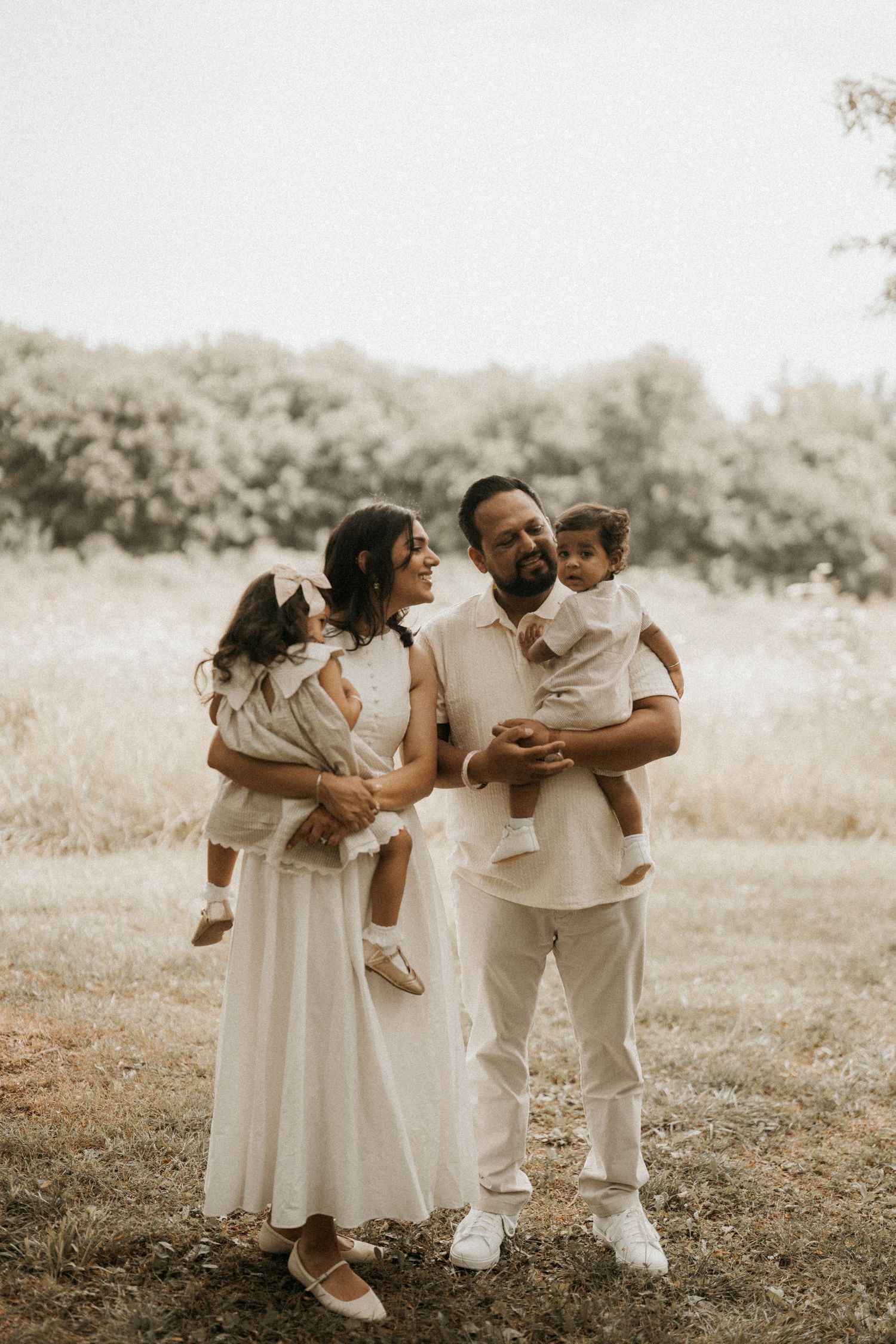 A happy family of four dressed in white poses outdoors in a natural field setting, parents each holding a young child.