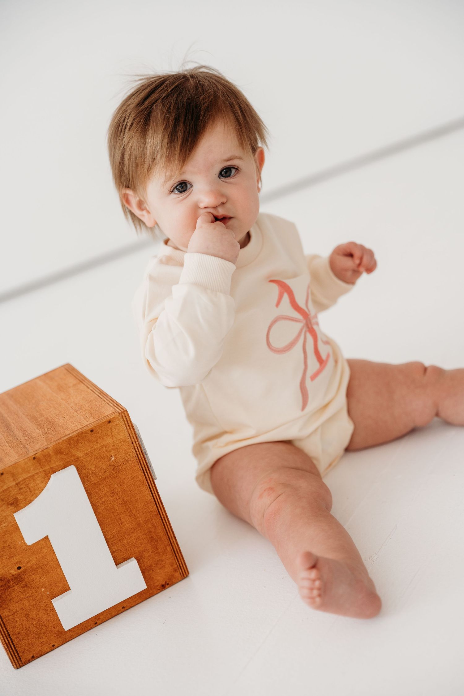 Baby in cream onesie with pink bow detail crawls on white floor near wooden number blocks.