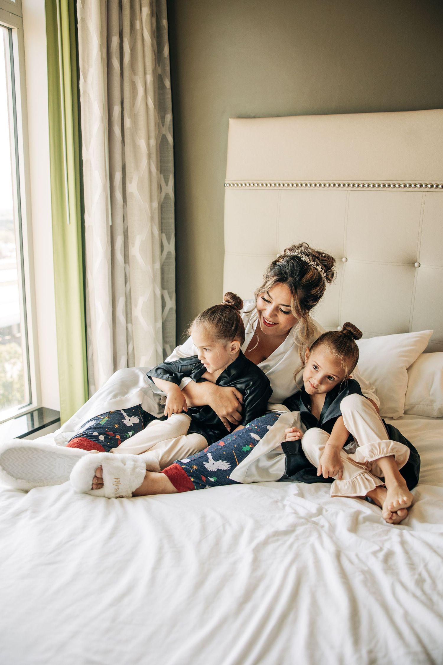 A family snuggles together under white blankets on a bed next to large windows with natural light streaming in.