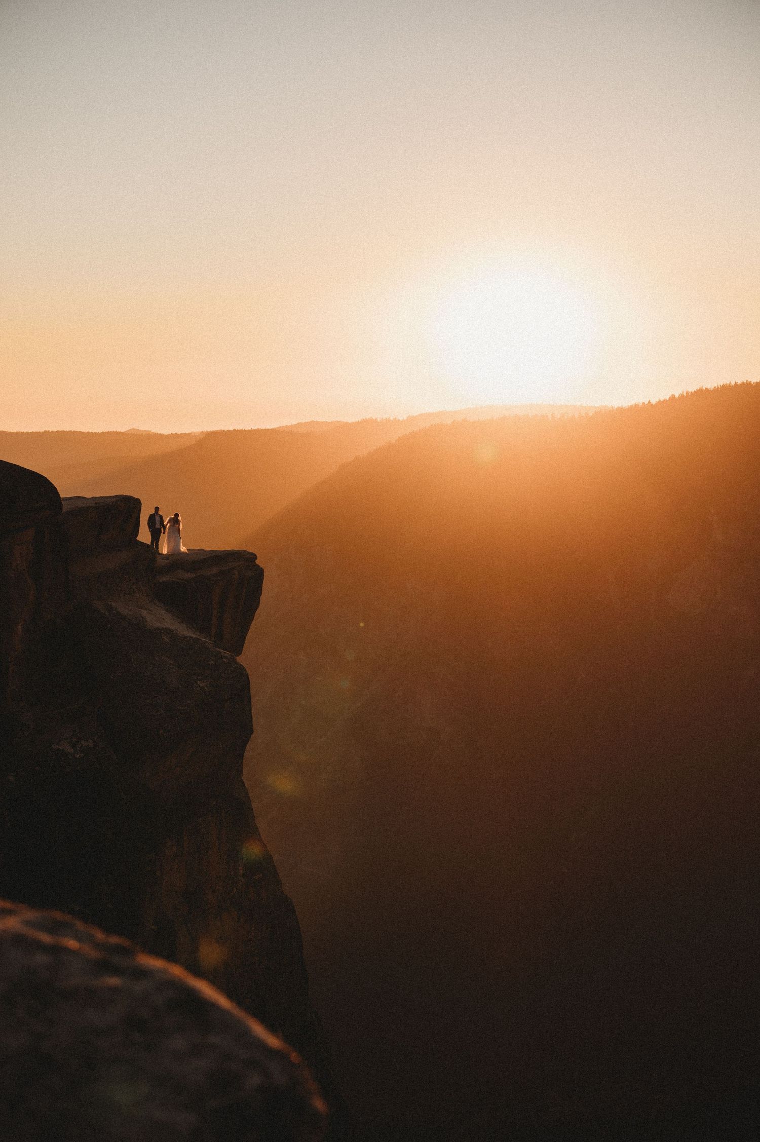 Silhouettes stand on a rocky cliff edge against a dramatic golden sunset over mountain ranges in the distance.