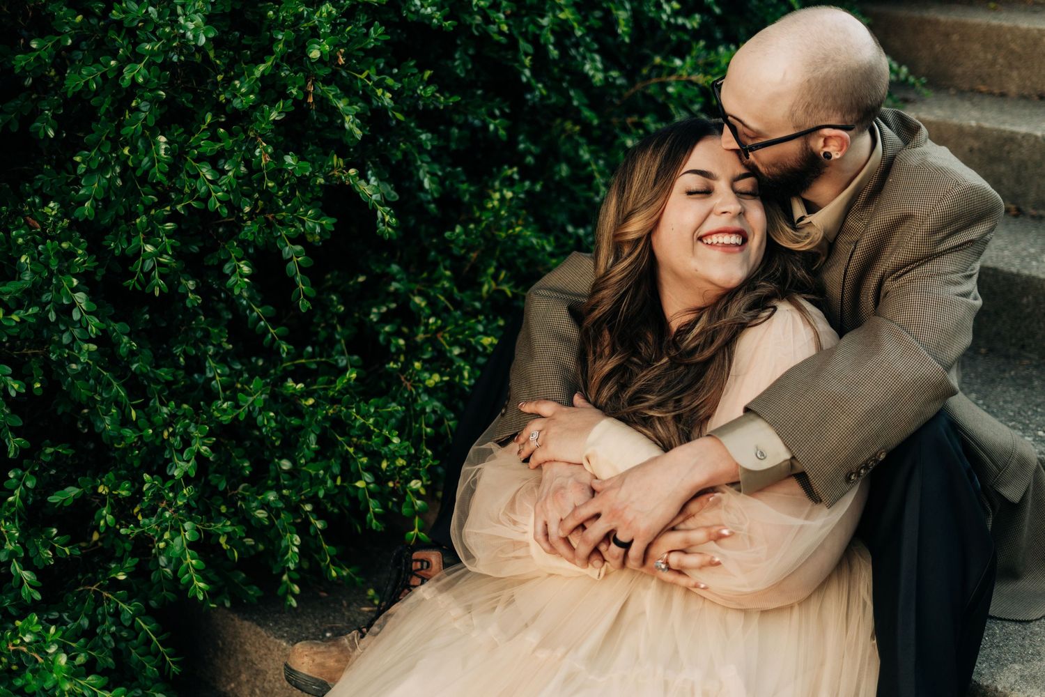 A couple embraces on steps next to lush greenery, with the woman in a light dress and the man in a tan jacket.