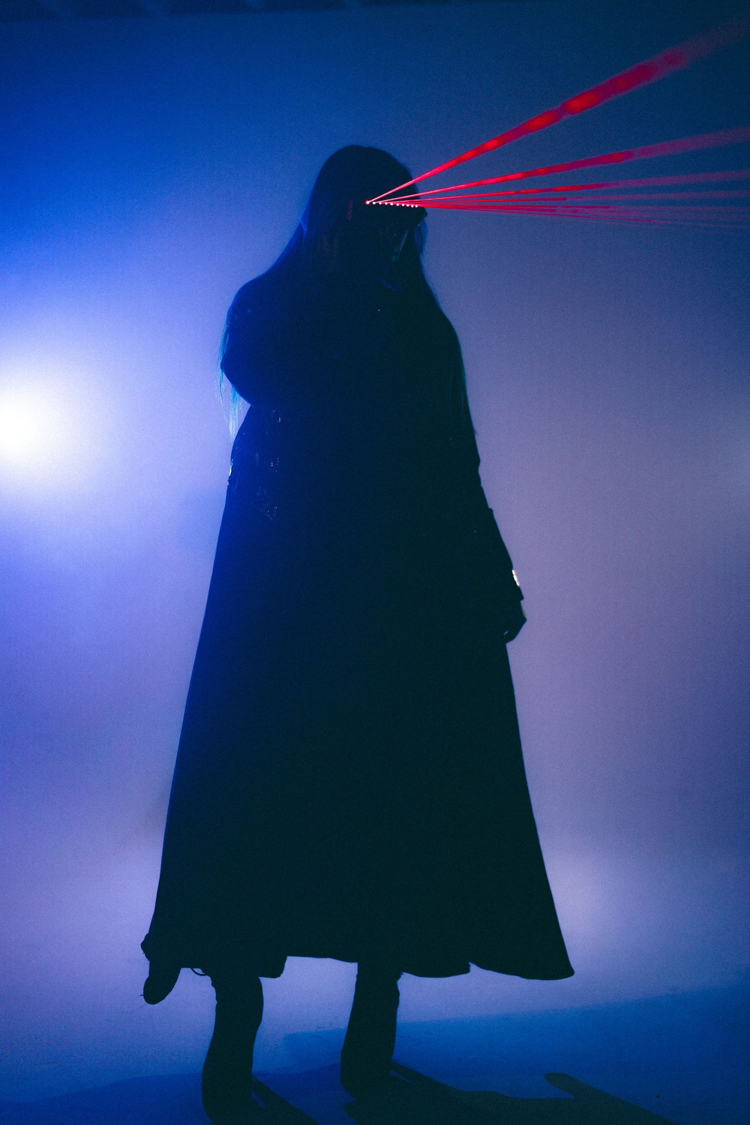 Silhouetted figure in long coat with glowing red laser beams shooting from eyes against dark blue atmospheric background.