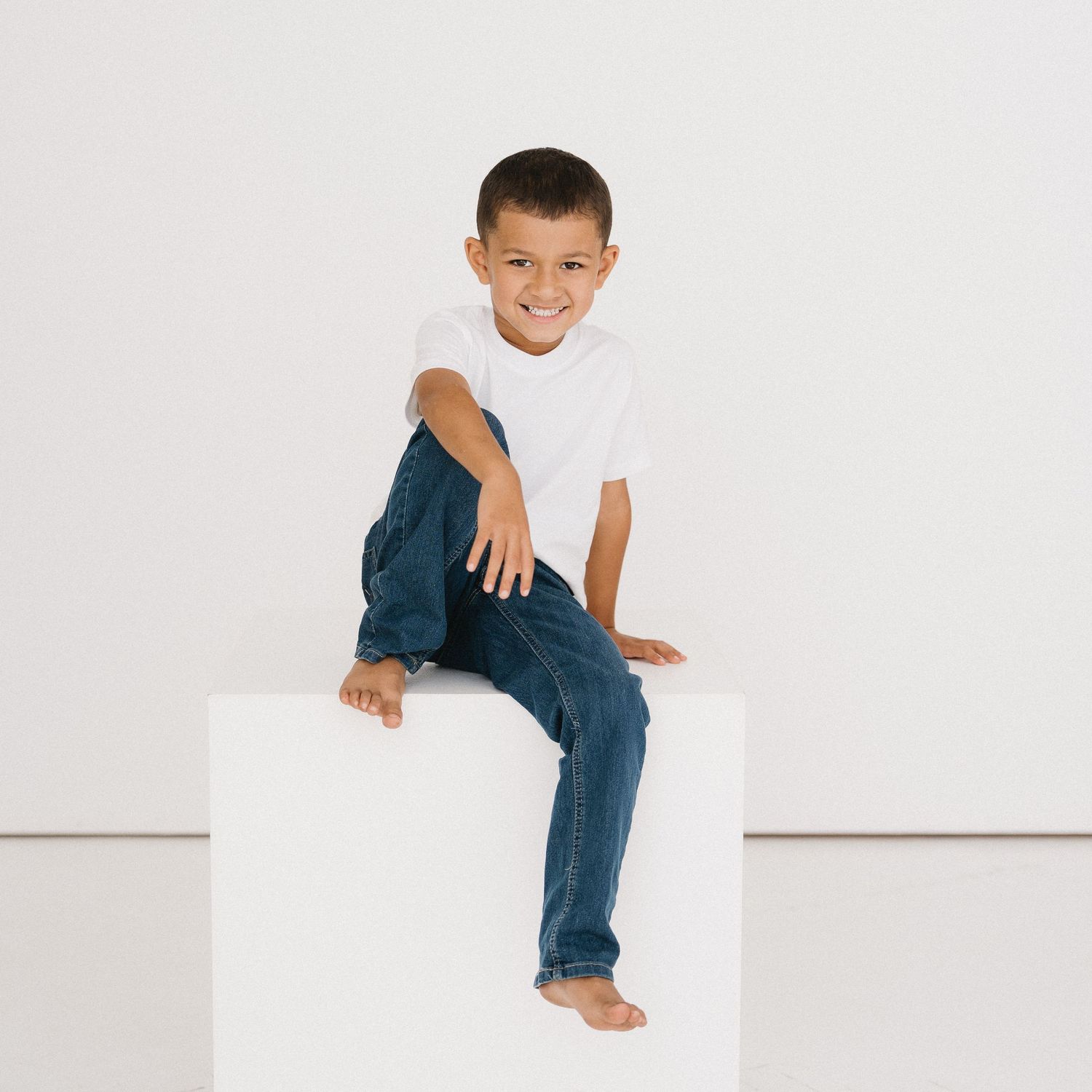 Child in white t-shirt and blue jeans sits casually on white platform against minimalist backdrop.