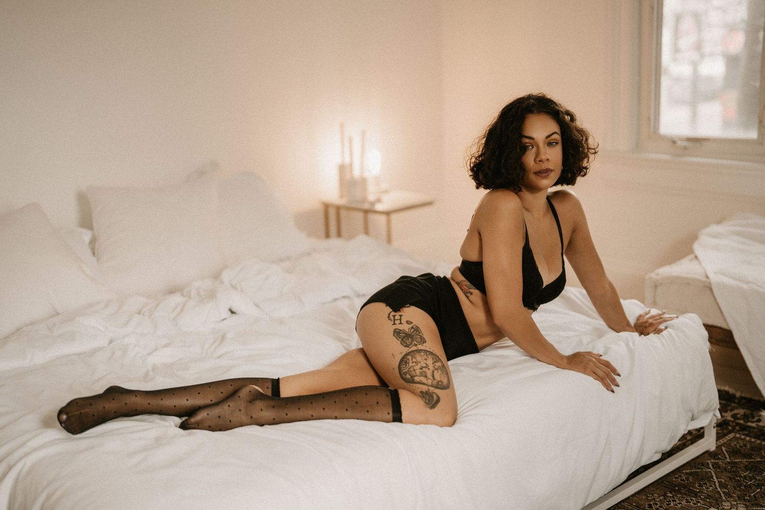 Woman in black lingerie and stockings posing on a white bed in a softly lit bedroom.