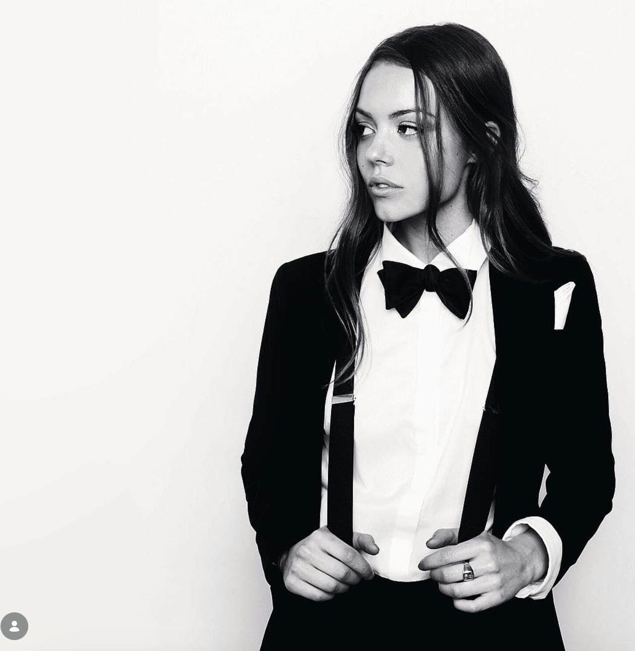 Black-and-white portrait of a woman with long dark hair in a tuxedo jacket, white shirt, bow tie and suspenders, gazing to the side.