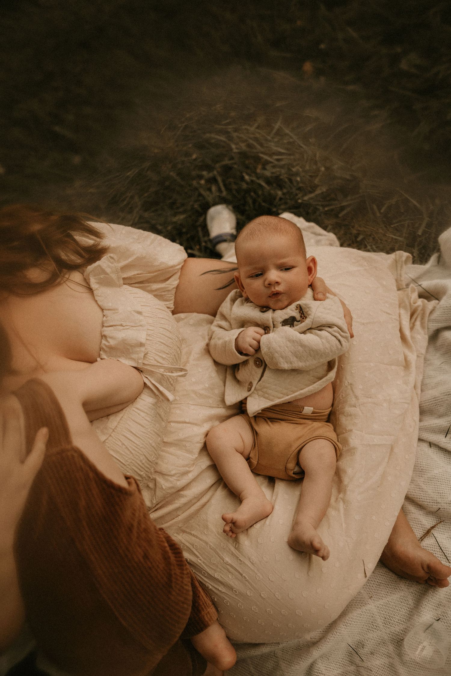 A newborn baby in a beige cardigan lies on a cream blanket beside a woman outdoors.